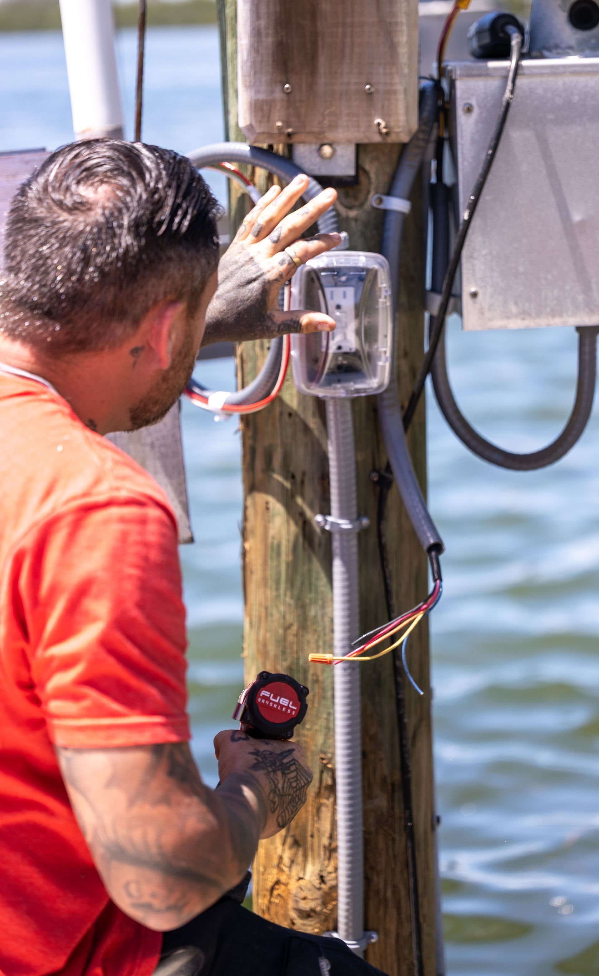 A person in a red shirt works on electrical wiring attached to a wooden pole near water.