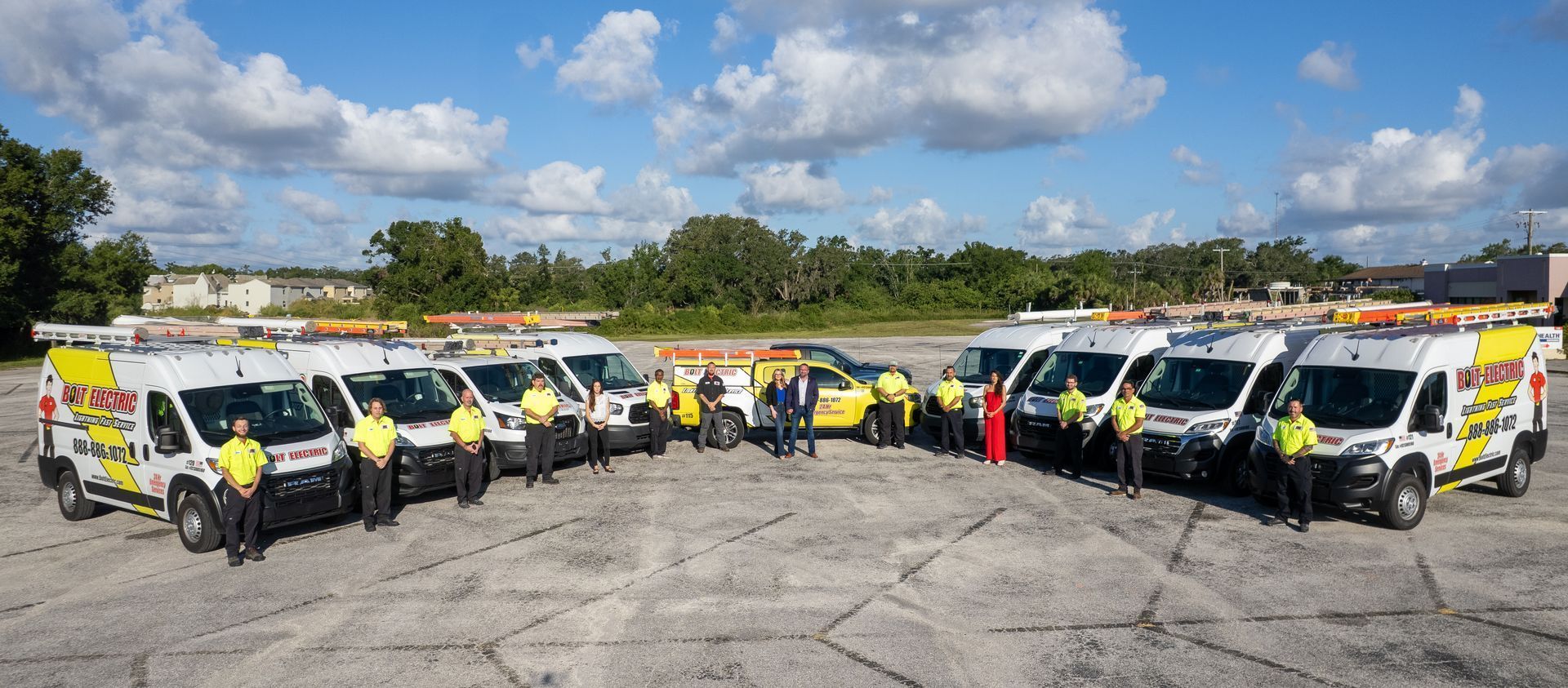 A group of people standing with service vans and a yellow car in a parking lot under a blue sky.