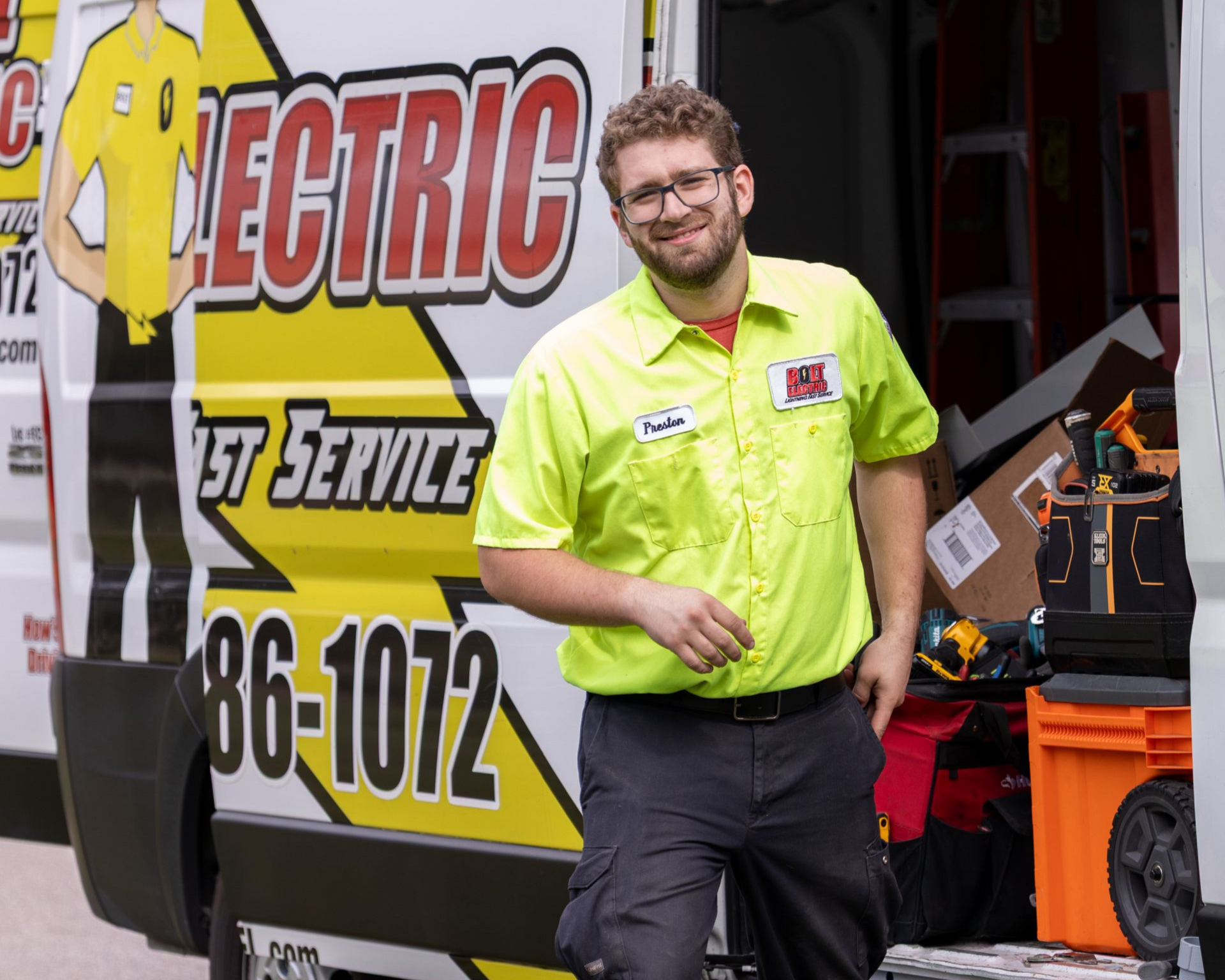 An electrician stands beside a van with an open door. He is smiling in front of a van that has tools and a ladder on top.