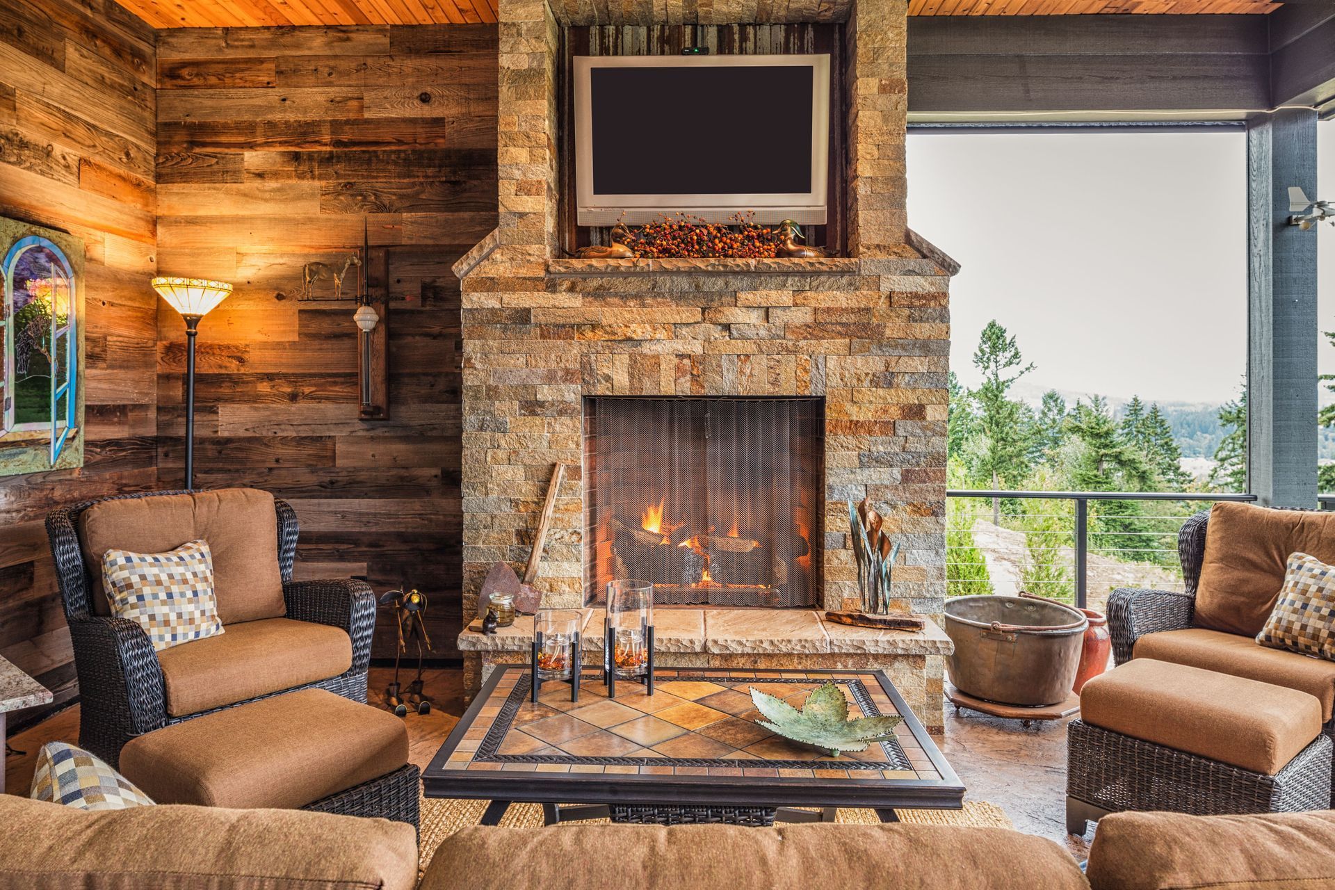 Outdoor living space with stone fireplace, TV, and wicker furniture overlooking trees.