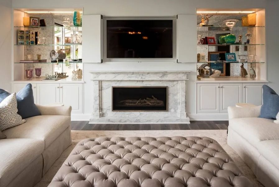 Living room with fireplace, built-in shelves, TV above, and tufted ottoman. Beige sofas flank the ottoman.