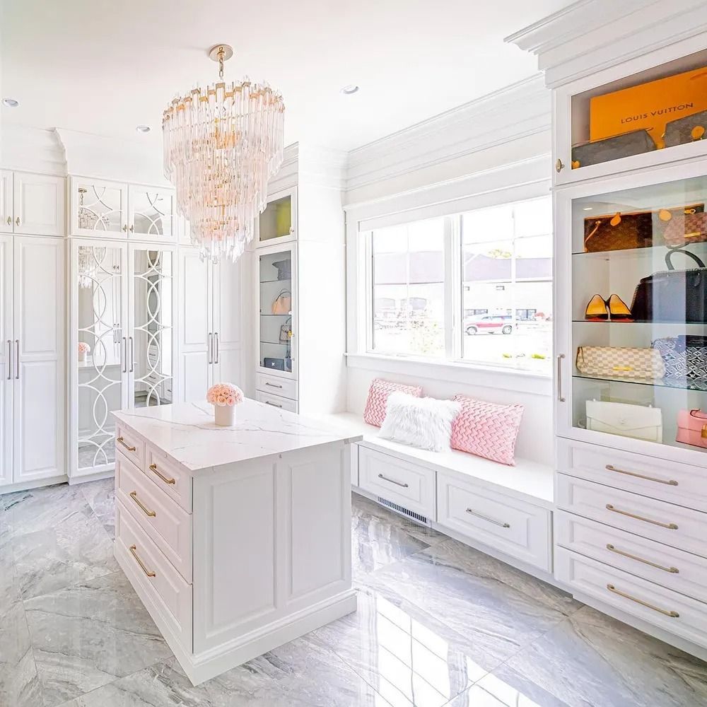 Elegant white closet with island, window seat, crystal chandelier, and marble floors.