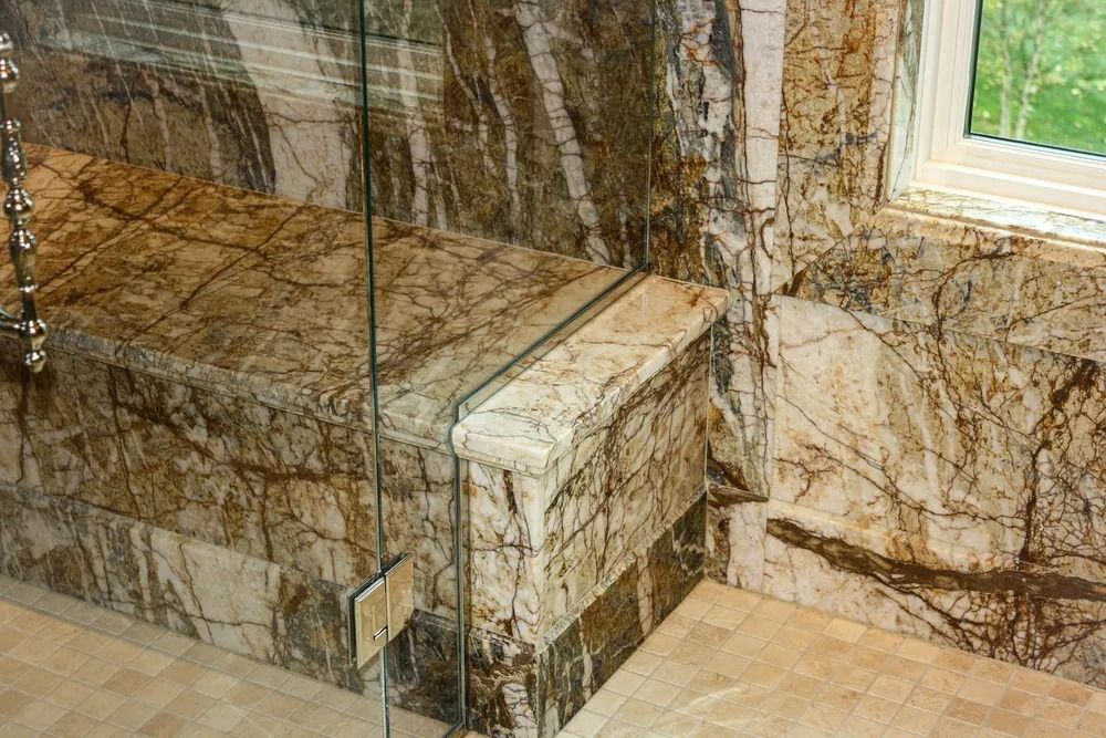 Shower with brown and white veined marble walls, corner bench, and glass door.