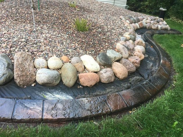 A curved landscape border with stamped concrete edging, topped by a row of river rocks and dark weed barrier fabric.