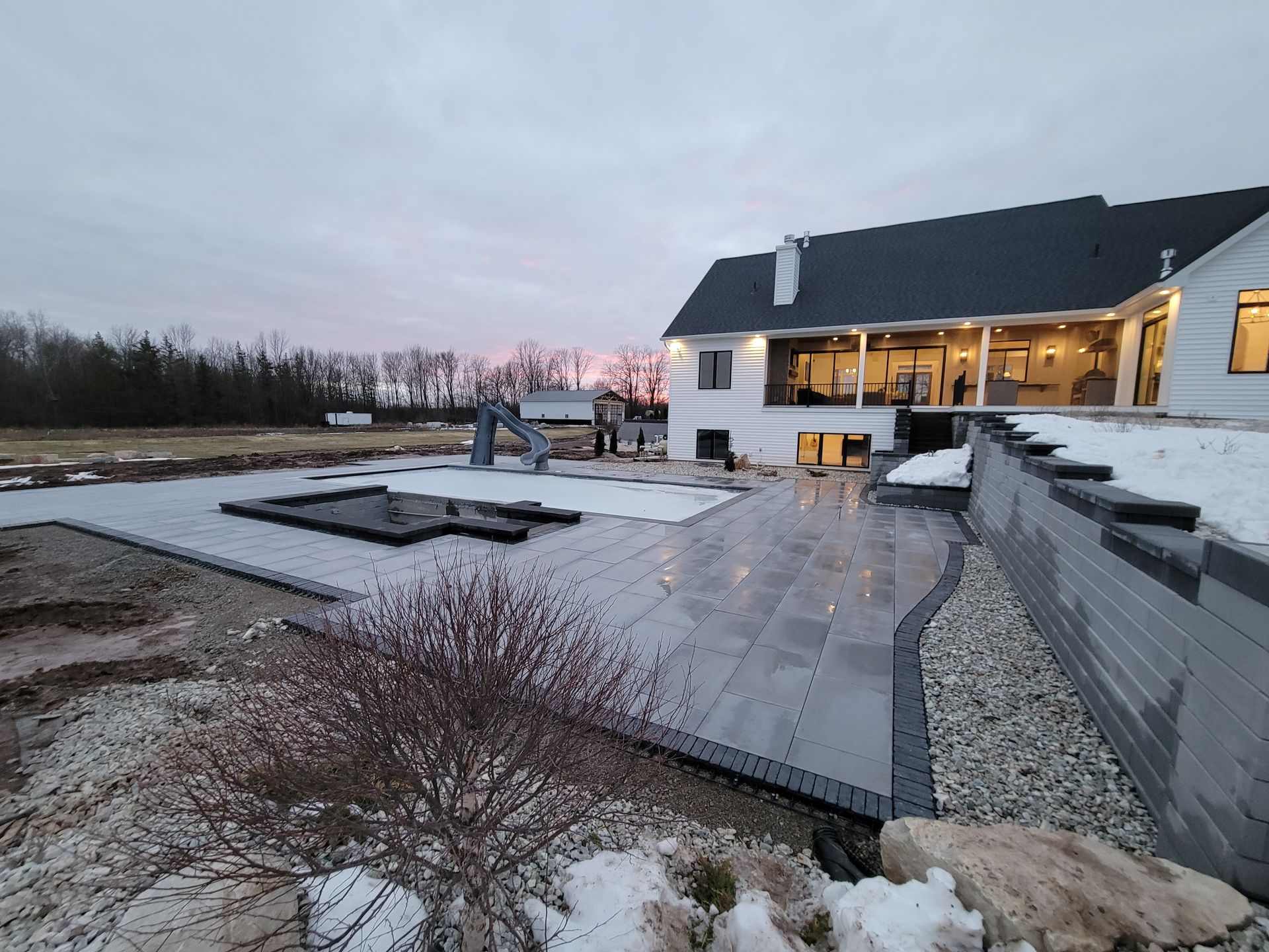 A modern house with a large paved patio and swimming pool during a snowy twilight.