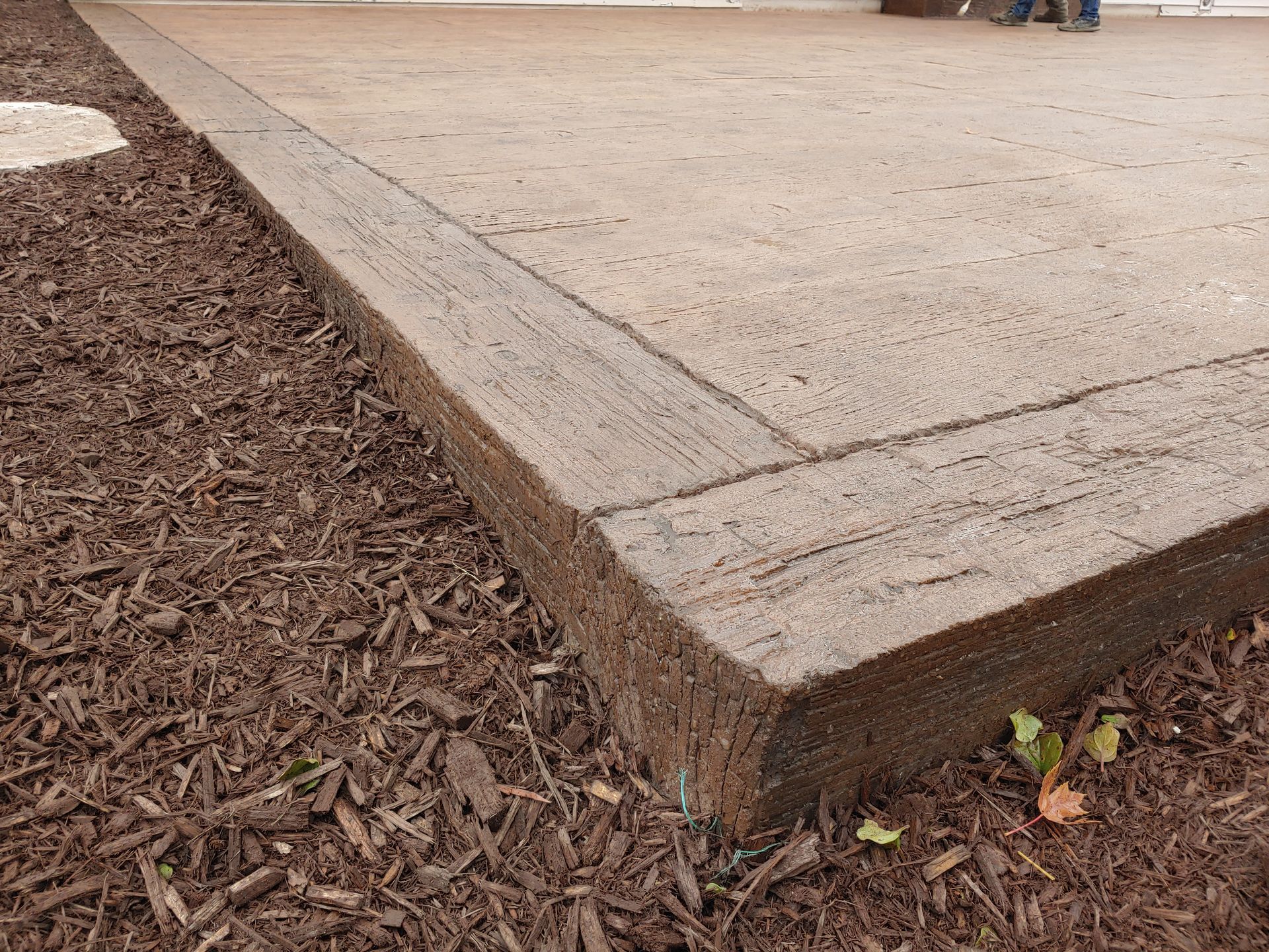 A textured, brown concrete patio edge bordered by a matching stamped trim, situated next to dark brown wood mulch.