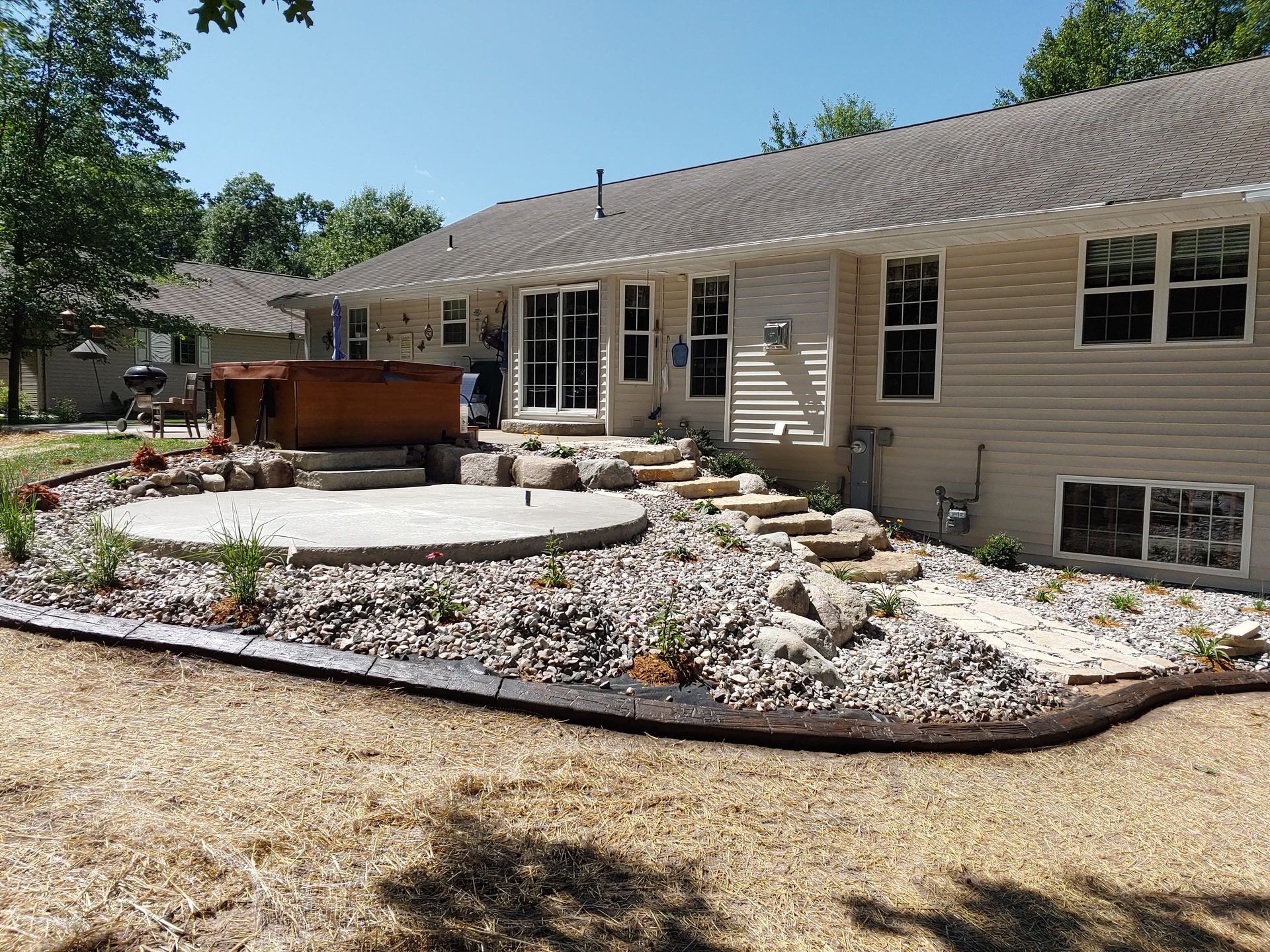A beige house features a backyard with a stone patio, a wooden hot tub, landscape rocks, and stone steps.