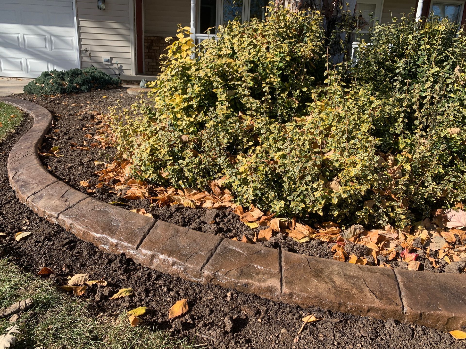 A decorative curved concrete landscape border separates a lawn from a shrub-filled garden bed with scattered leaves.