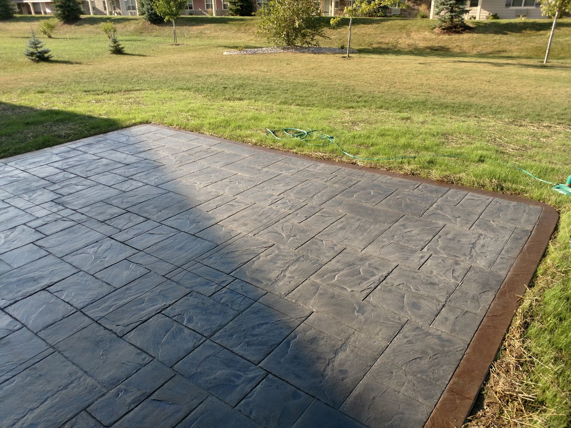 Gray stamped concrete patio with a rectangular pattern and a dark brown border set in a grassy backyard.