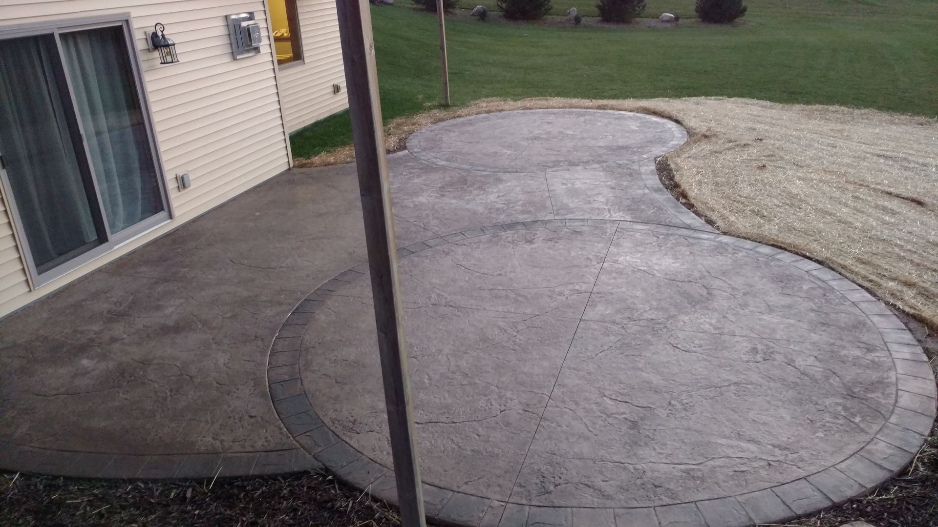 A stamped concrete patio with circular designs and a dark border, located next to a house with beige siding.