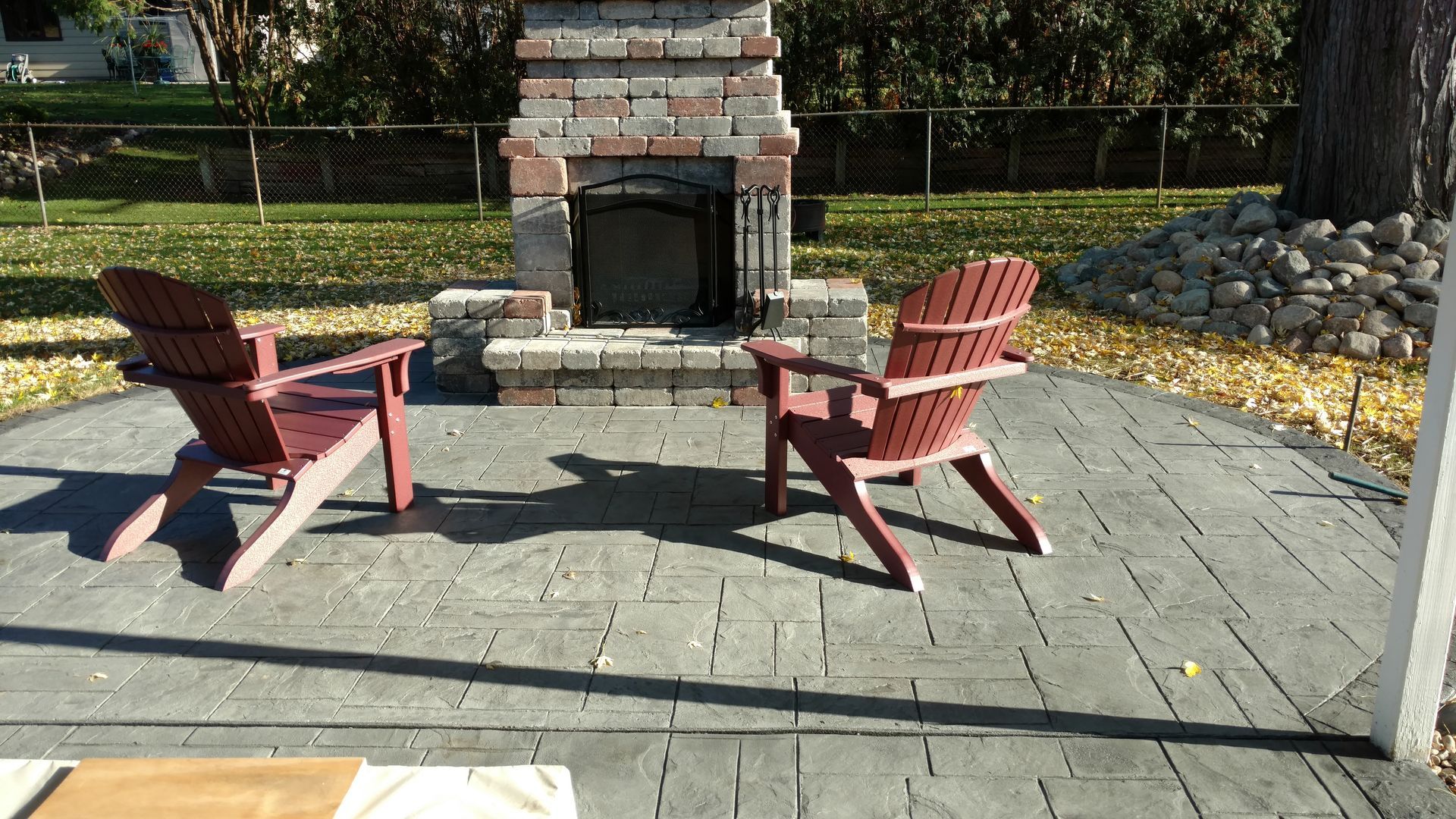 Two red Adirondack chairs face a brick outdoor fireplace on a grey paved patio in a grassy yard.