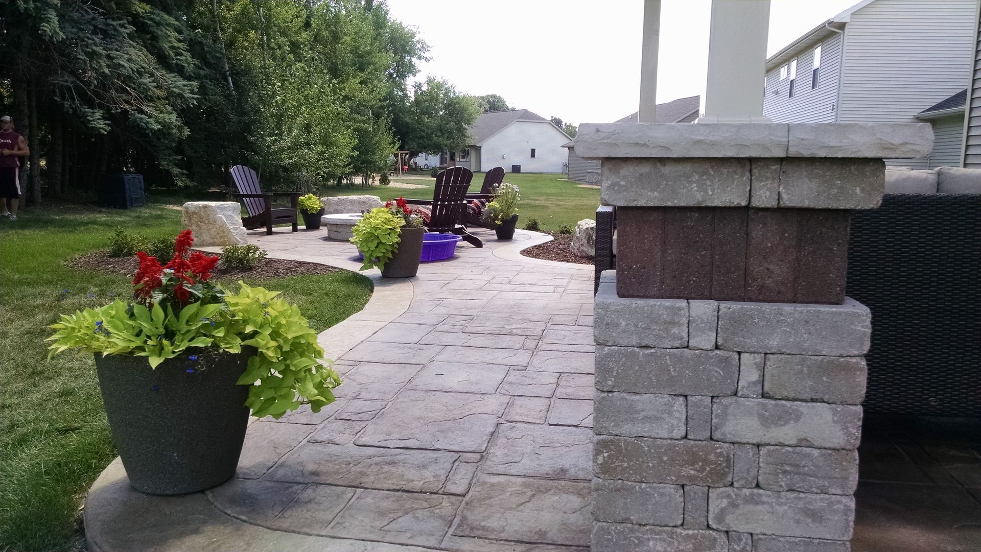 A stone patio with a decorative brick column, potted plants, and outdoor furniture in a backyard setting.