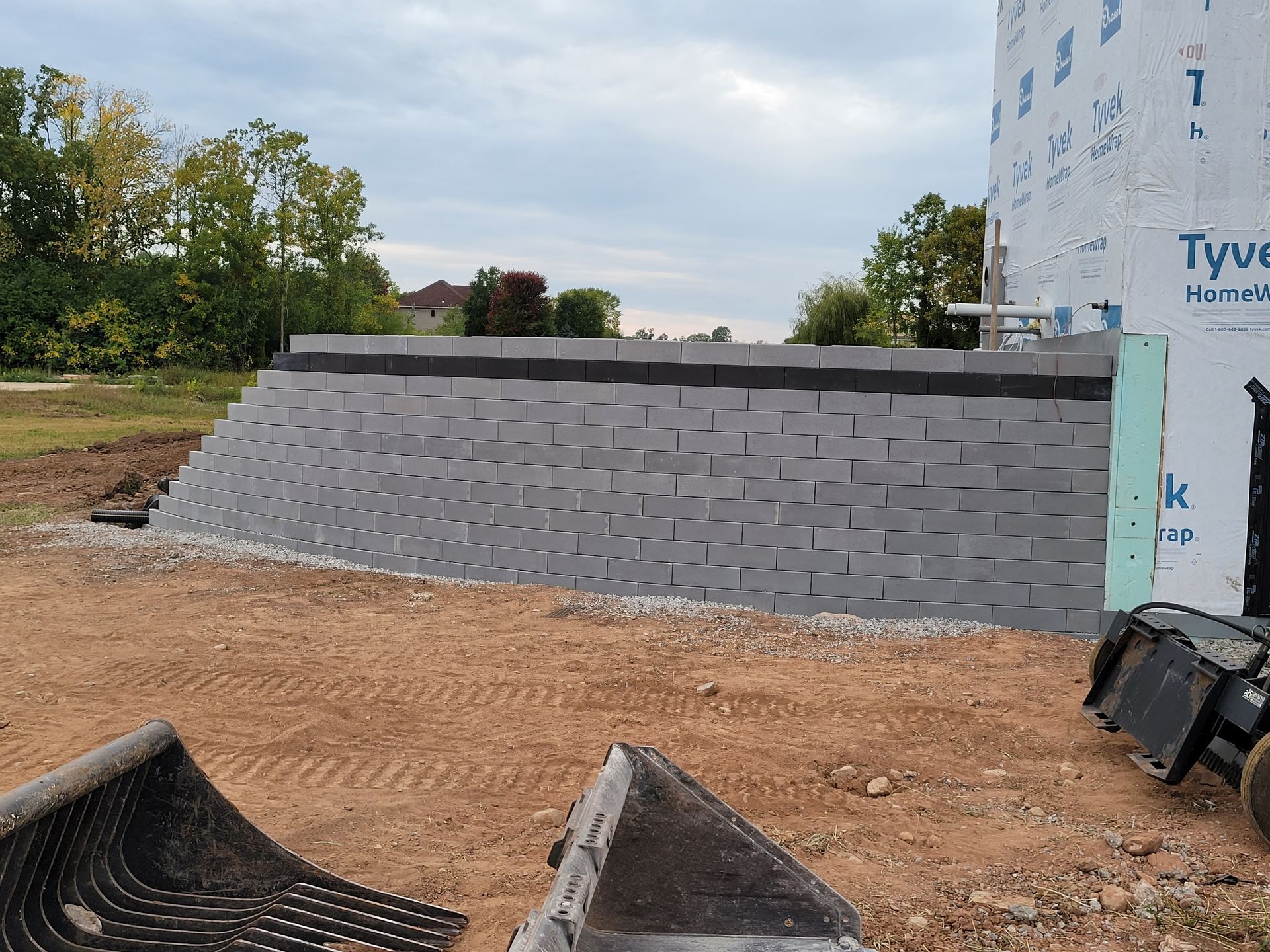 A gray concrete block retaining wall under construction next to a building covered in Tyvek wrap at a dirt work site.