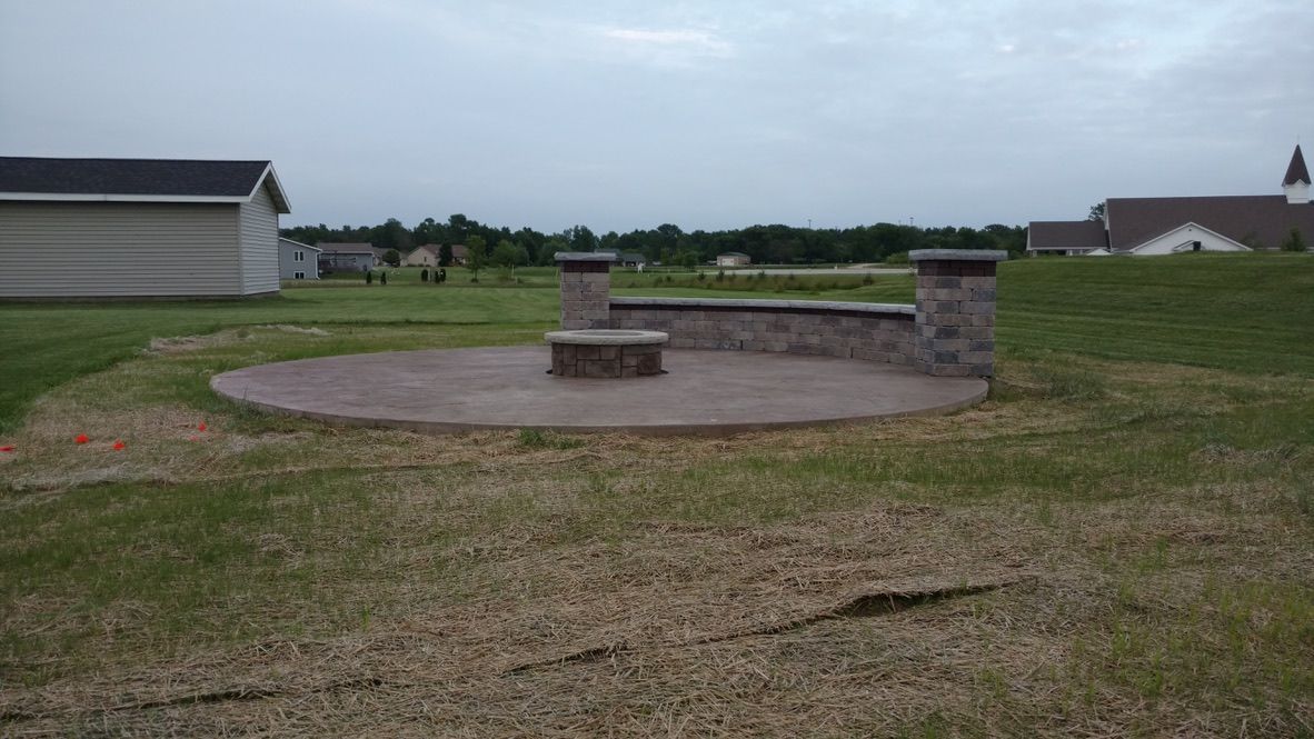 A circular stone patio features a built-in fire pit and a curved seating wall with two stone pillars in a grassy yard.