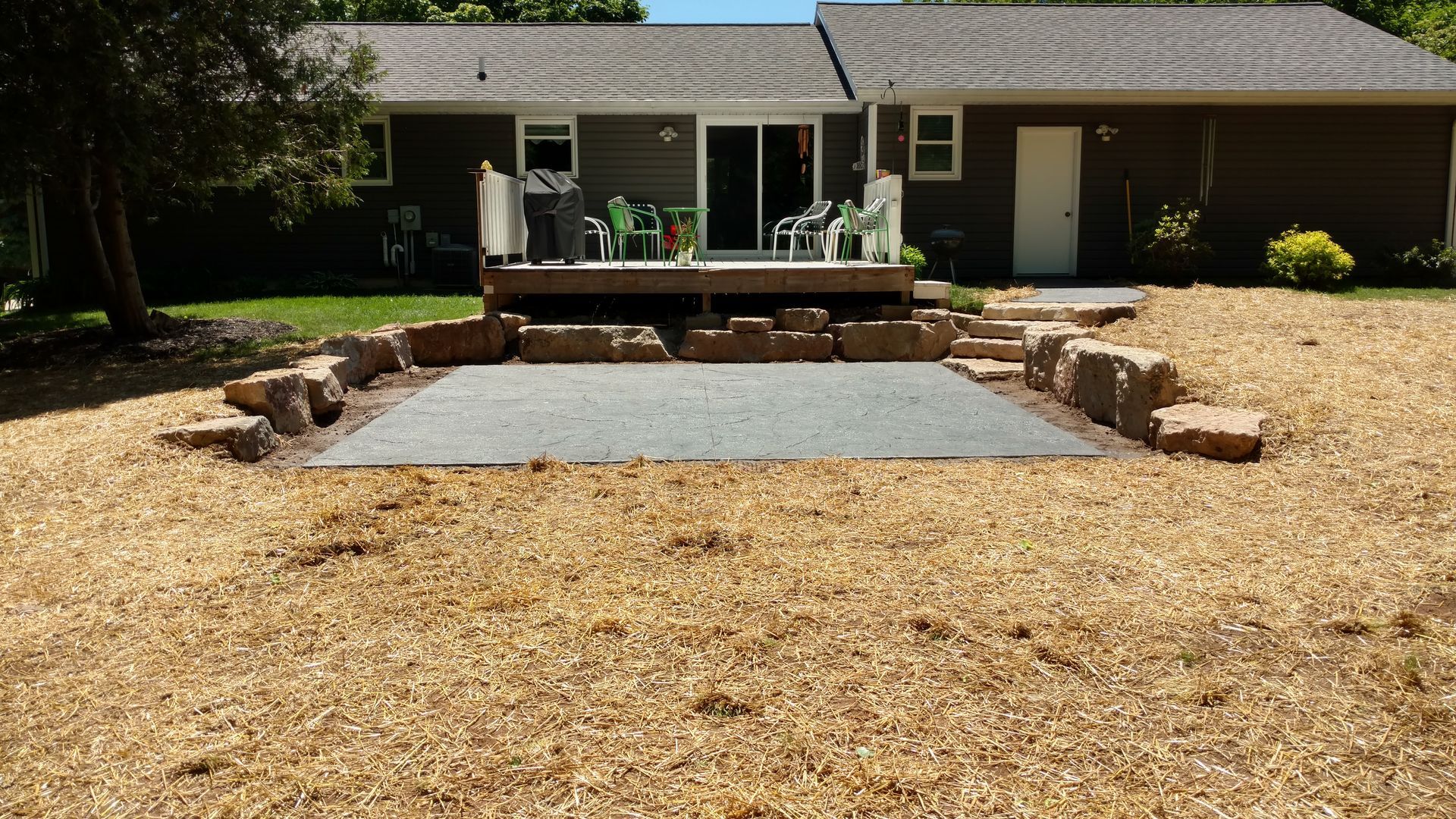 A gray gravel patio area surrounded by large stone blocks, set in a backyard with wood chips and a wooden deck.