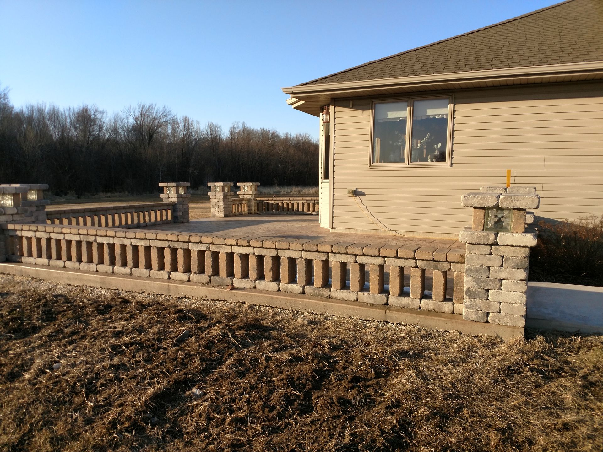 A tan house with a stone patio featuring a decorative brick railing and pillars in a rural, grassy setting.