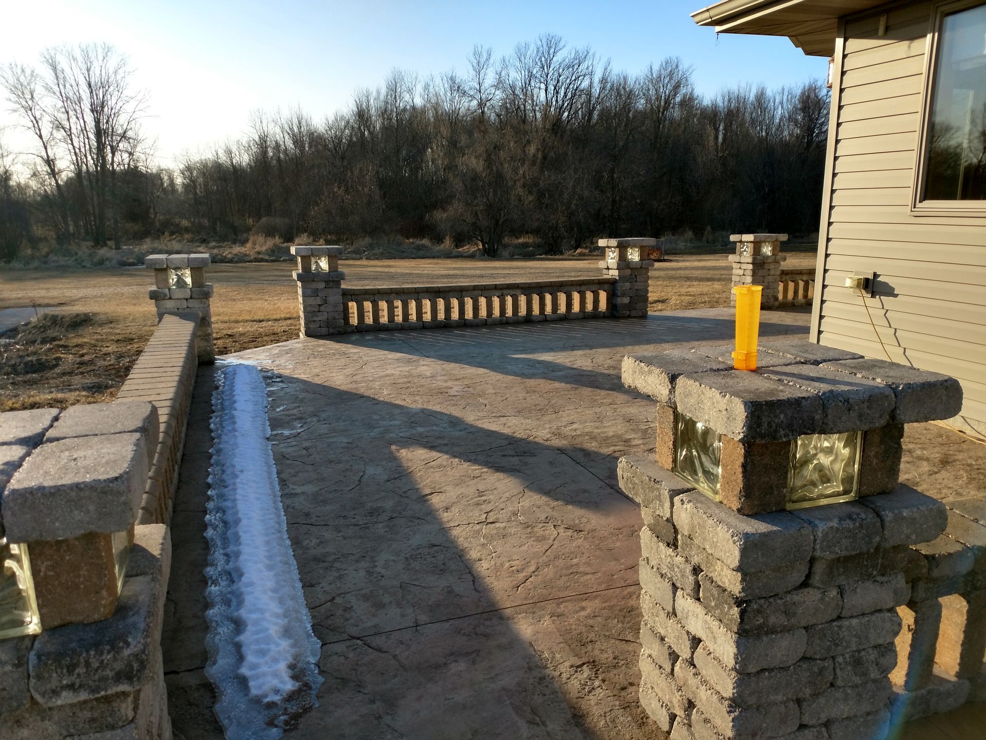 A stone patio with a balustrade, pillars with glass blocks, and a patch of snow, beside a tan house under a clear sky.