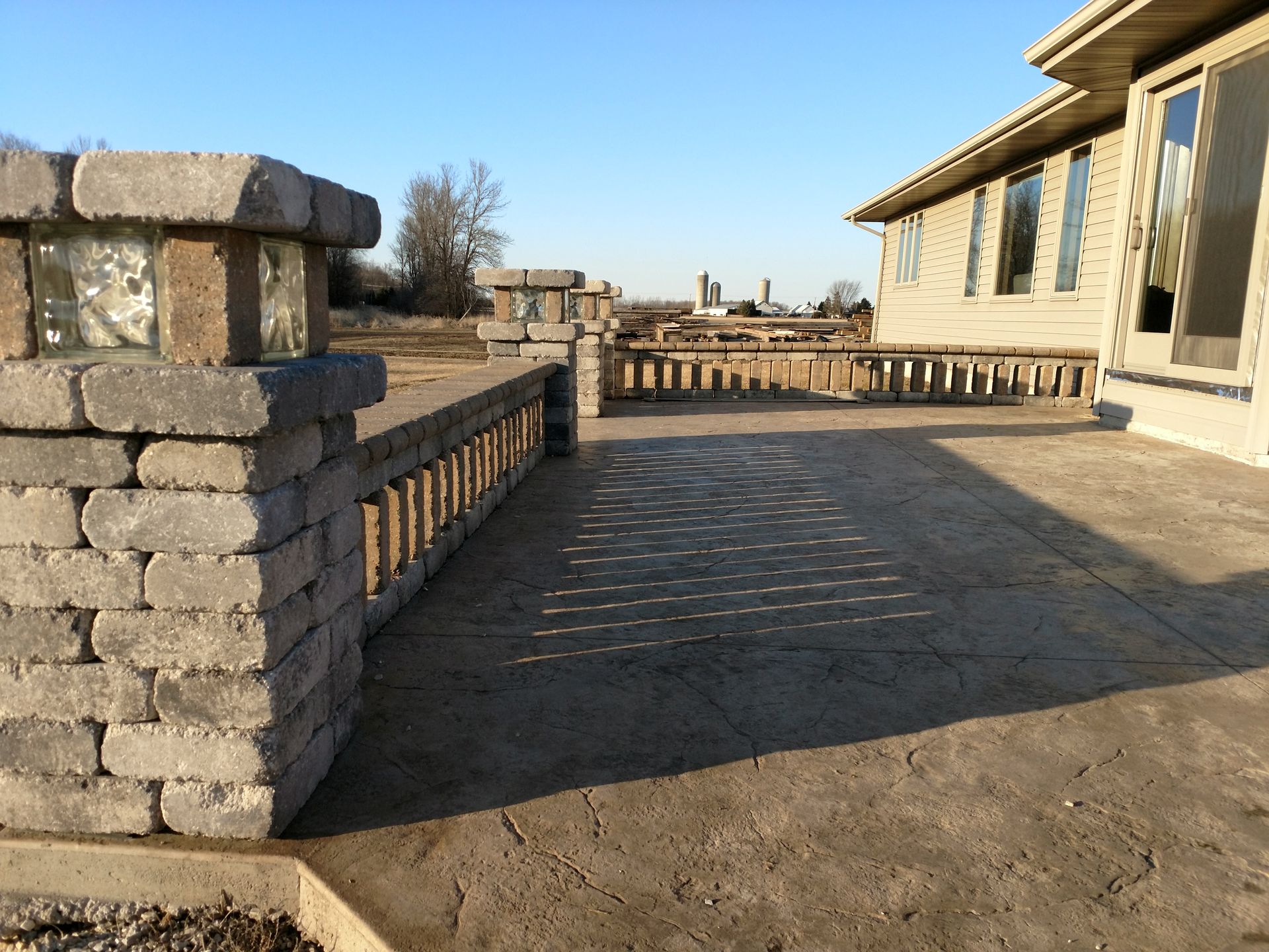 A stone patio features a low wall with illuminated glass block pillars, adjacent to a house exterior under a clear sky.