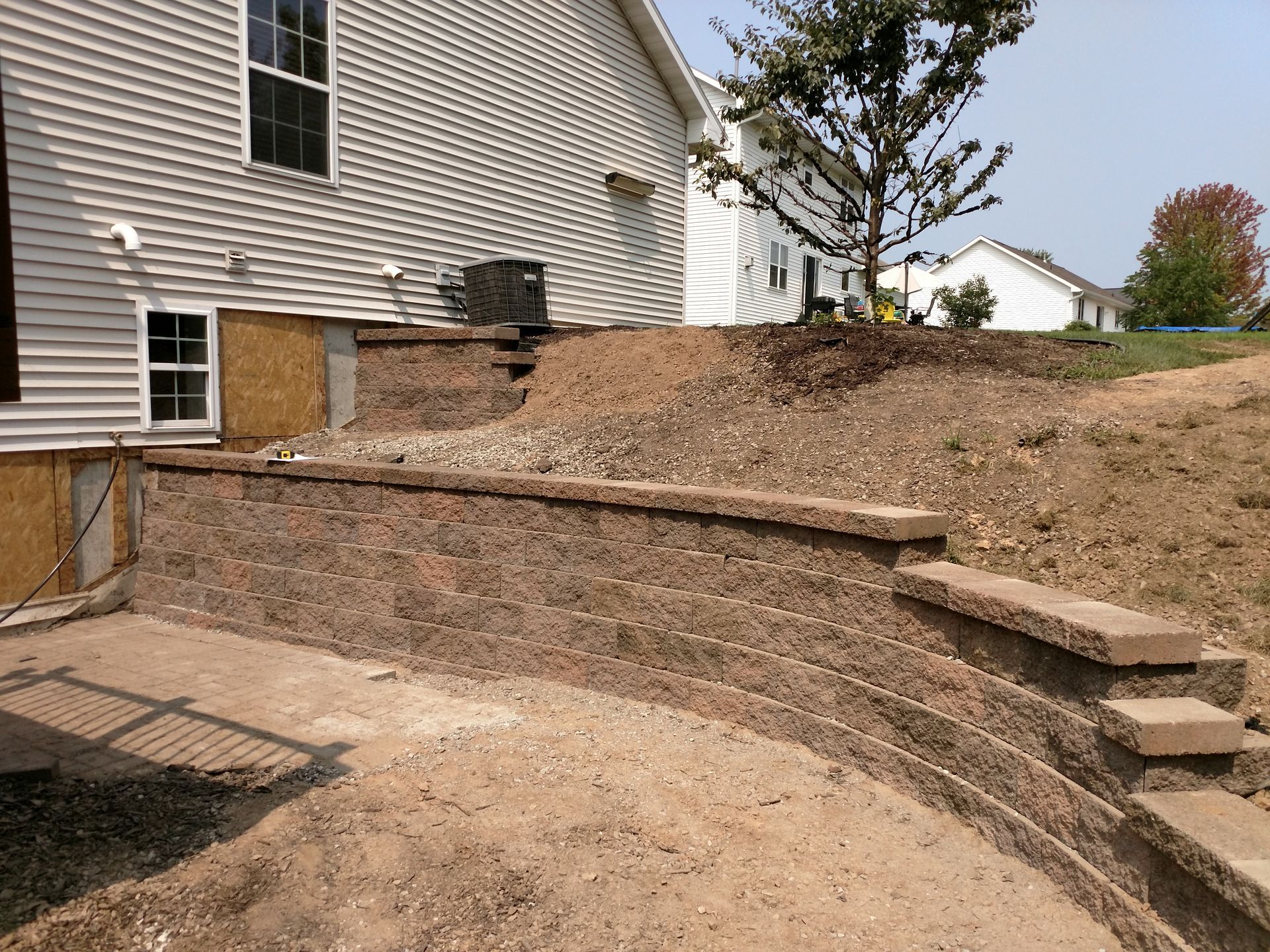 A newly constructed curved stone retaining wall separates a beige house from a sloped, unfinished dirt yard.