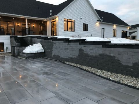 A modern house with a large gray paver patio and a tiered gray concrete block retaining wall, featuring patches of snow.