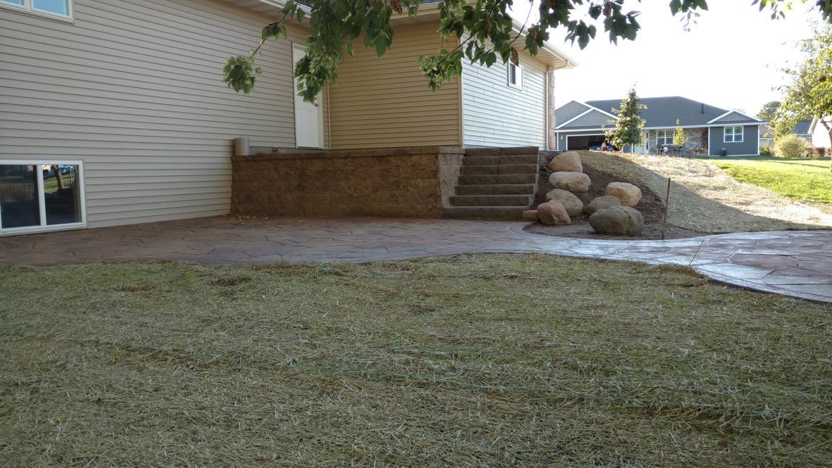 A patio and retaining wall with stone steps leading up to the side of a tan vinyl-sided house in a suburban backyard.