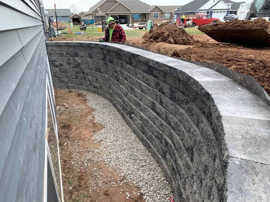 A construction worker in a high-visibility vest builds a curved stone retaining wall alongside a house foundation.