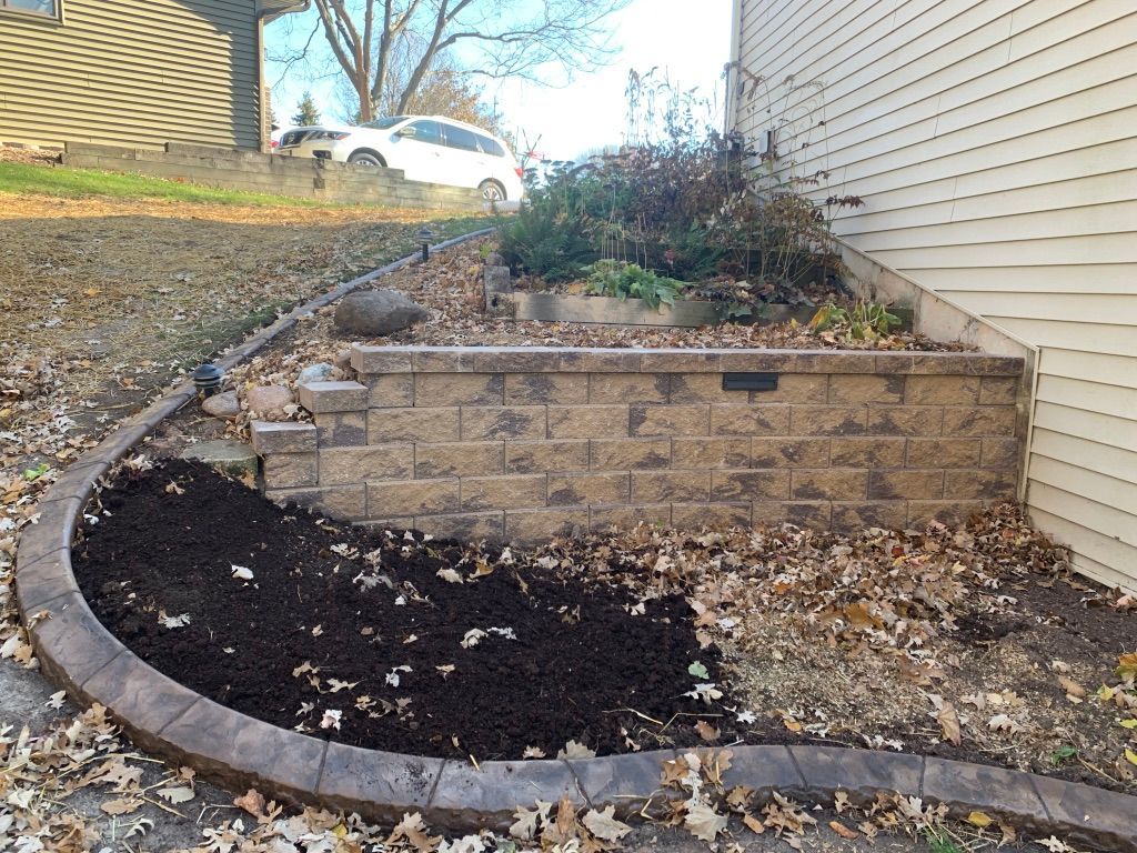 A curved retaining wall made of tan stone blocks borders a patch of fresh dark soil and fallen leaves next to a house.