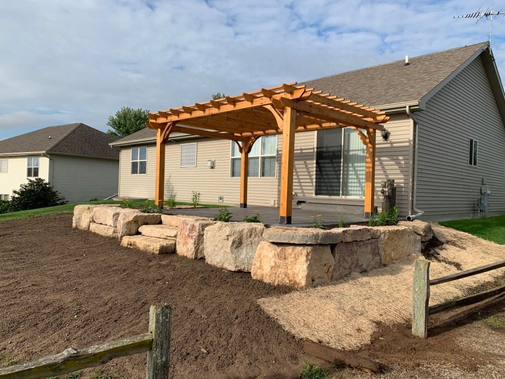 A tan house with a wood pergola on a stone-retained patio, overlooking a backyard with a split-rail fence.