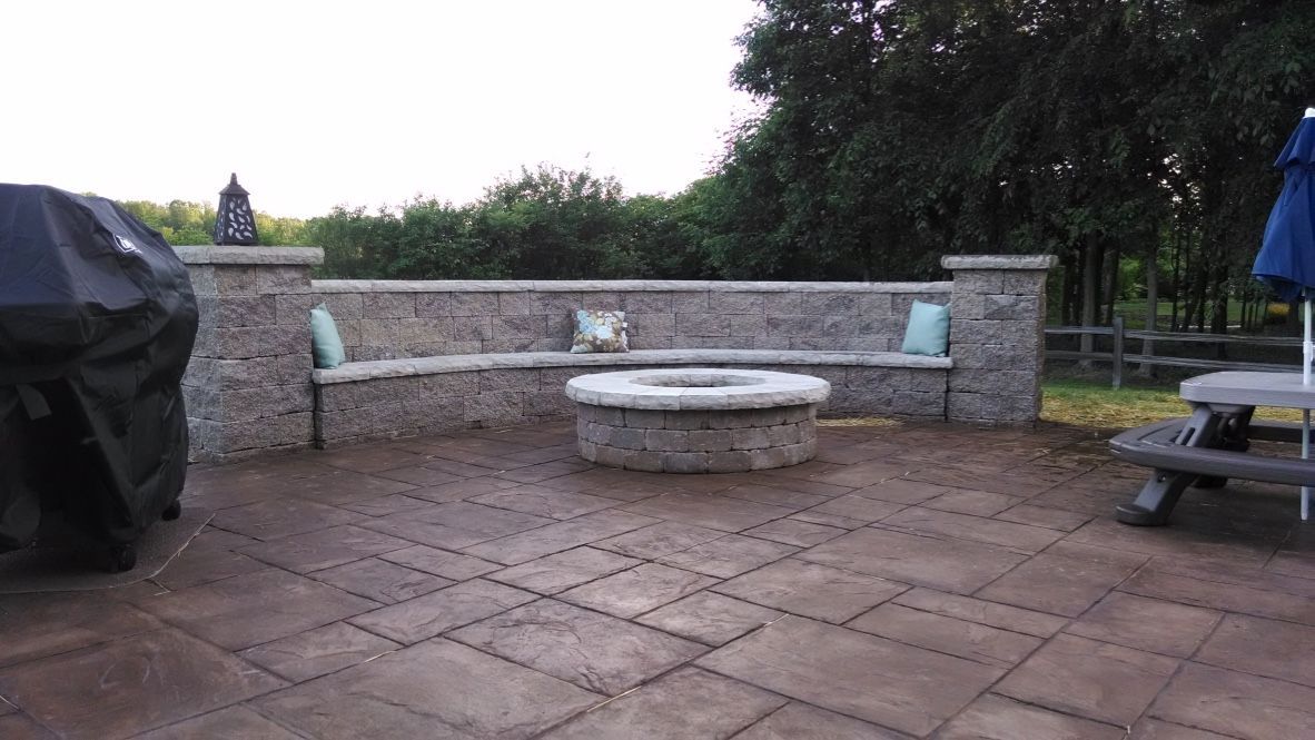 A stone patio with a curved built-in bench around a fire pit, next to a grill and a small outdoor table.