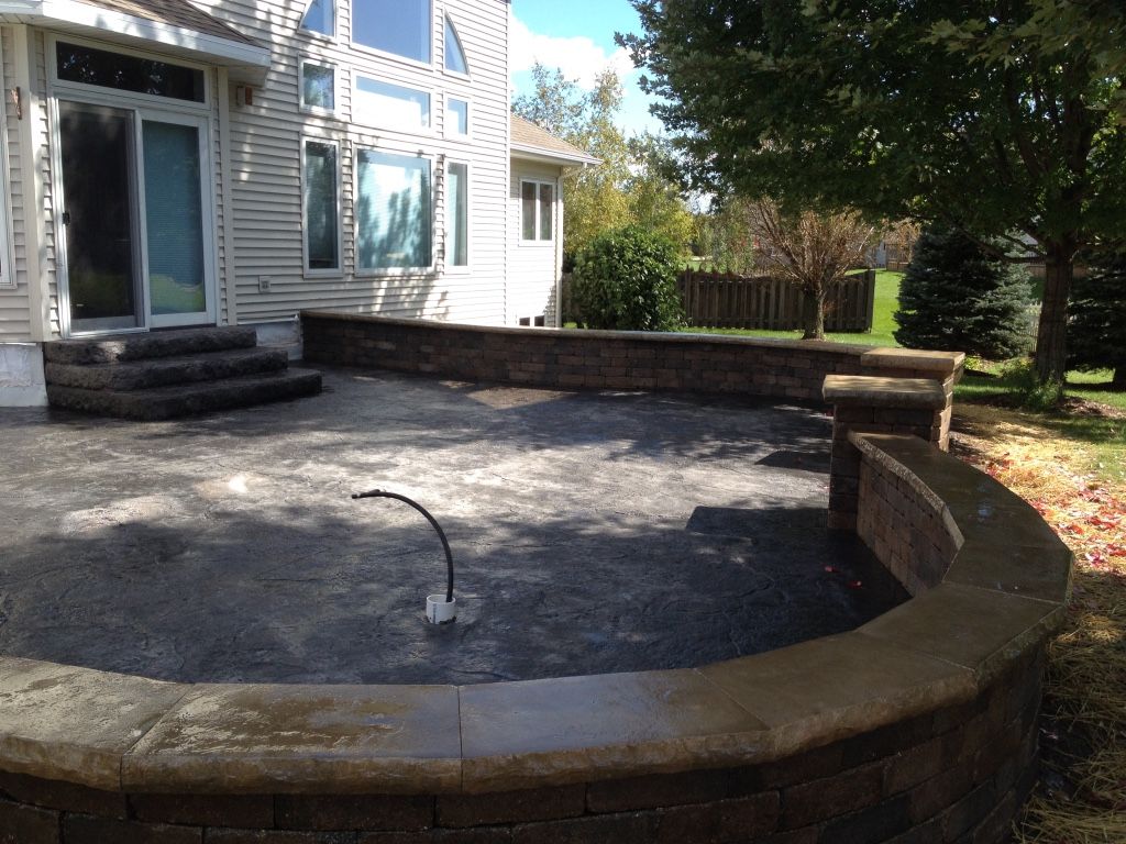 A stamped concrete patio featuring a curved retaining wall and stone steps leading to a house with beige siding.