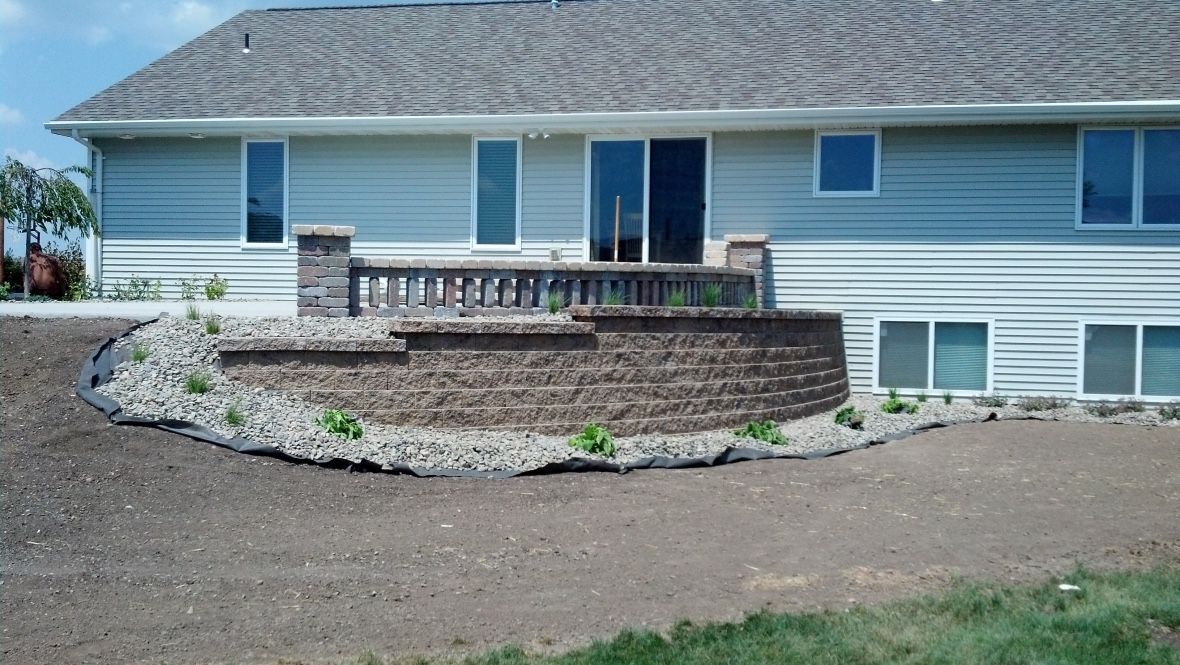 A beige house features a tiered, curved stone retaining wall patio with a decorative railing, set against bare dirt.
