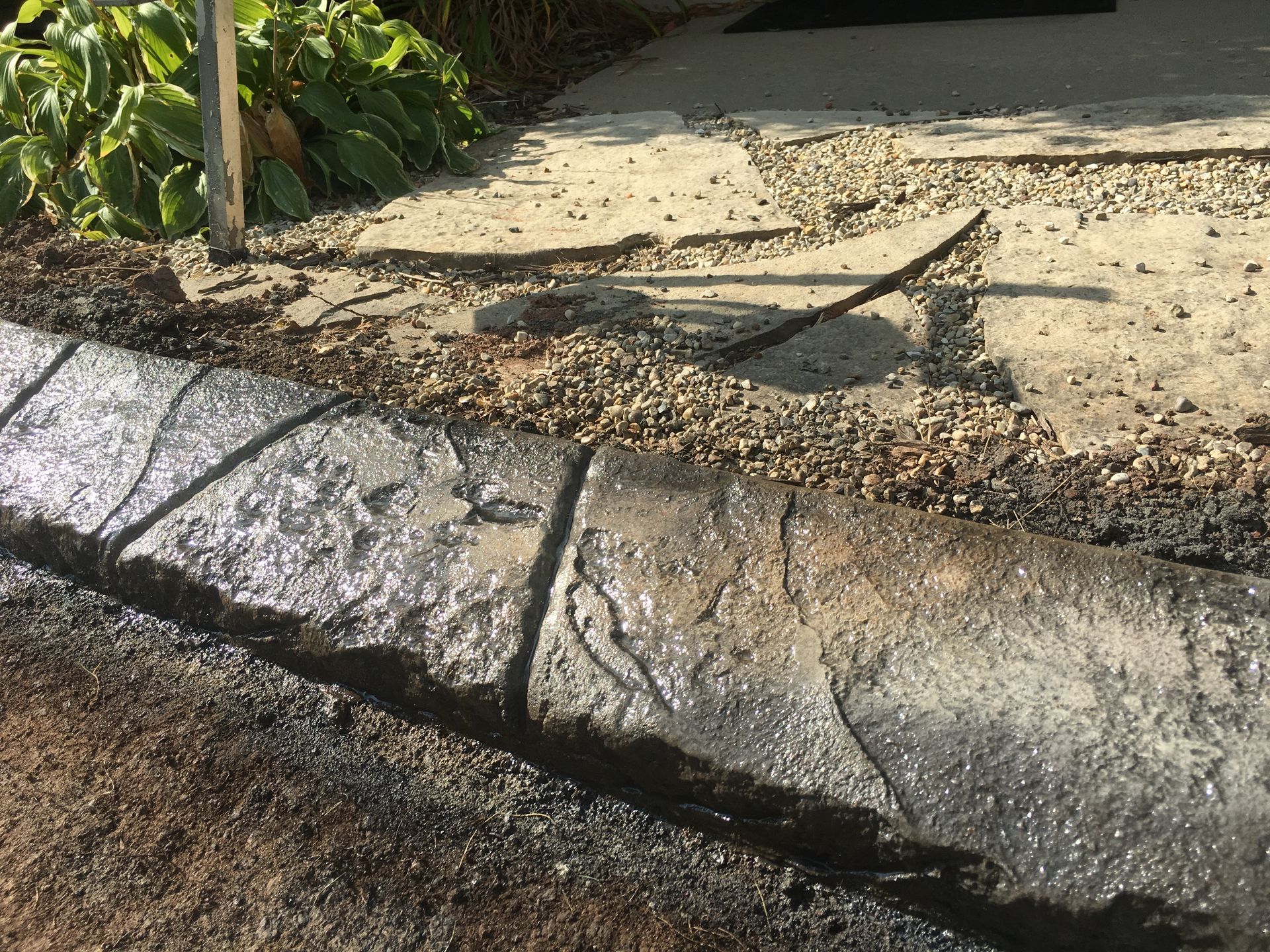 A textured, gray concrete landscape edging border sits between a patch of dirt and a graveled patio area.