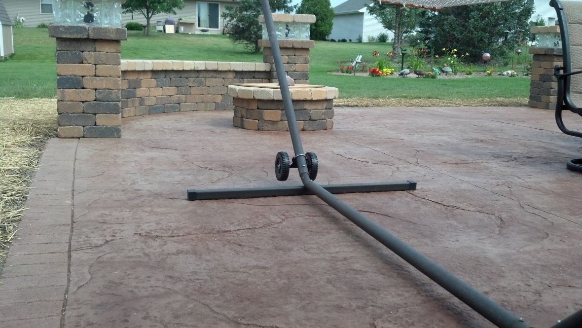 A black metal hammock stand rests on a textured, reddish-brown concrete patio in front of a stone fire pit and wall.