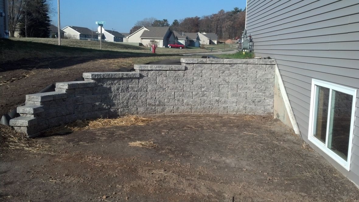 A grey stone retaining wall tiered down to meet the siding and white-framed basement window of a house.
