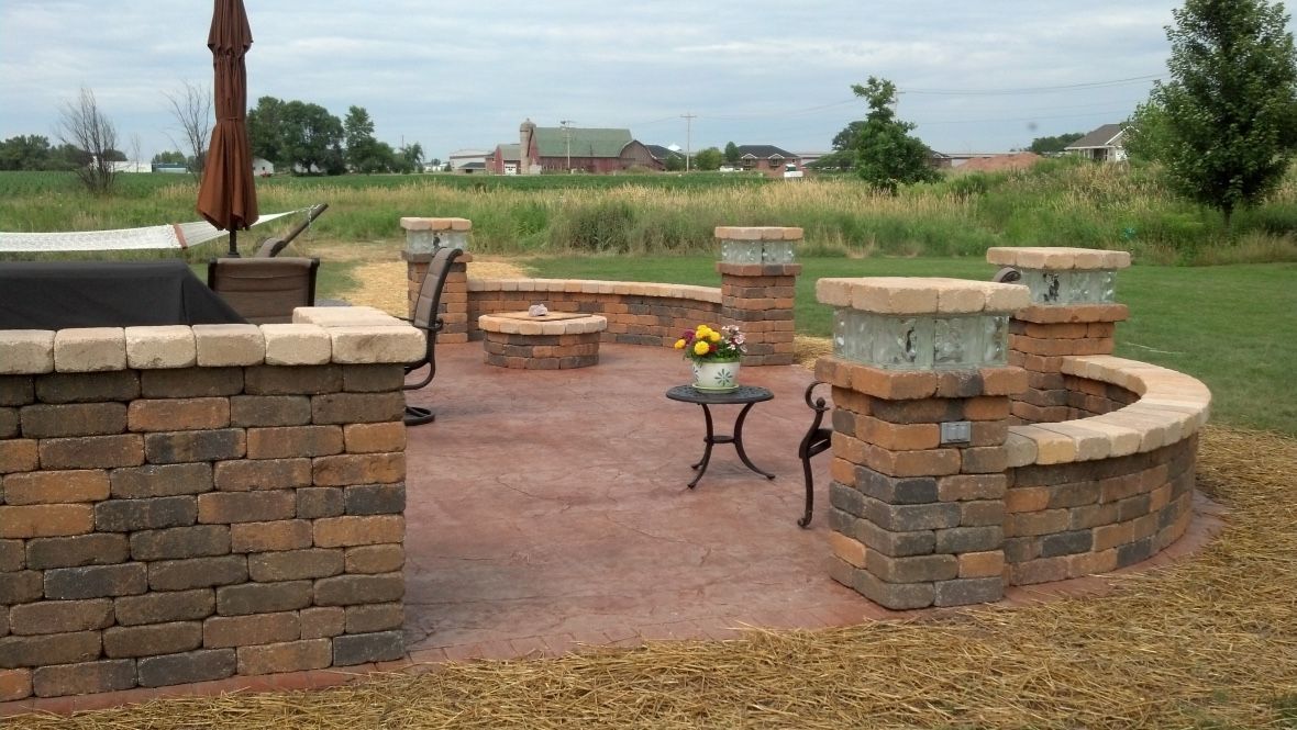 A backyard patio with a stone fire pit, seating wall, and pillars, set on a reddish-brown stamped concrete surface.