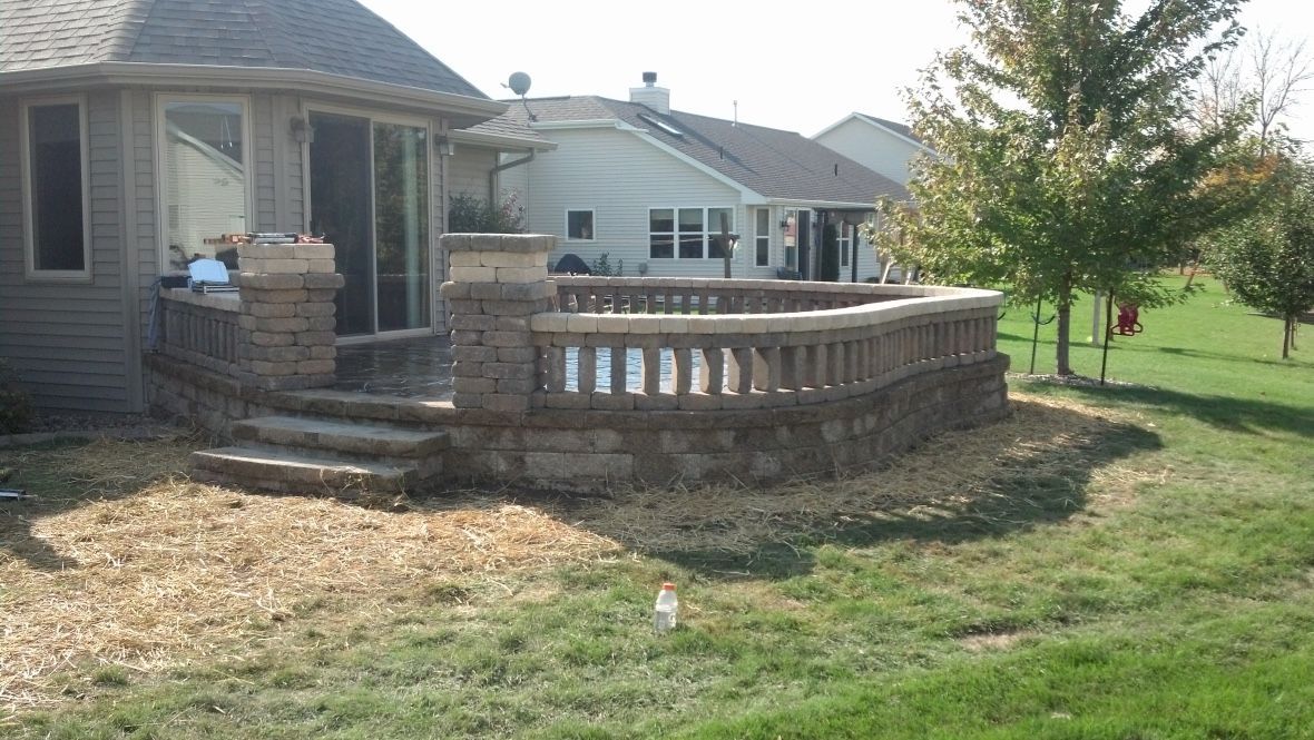 A stone patio with a balustrade railing attached to the back of a house, surrounded by a lawn with wood mulch.