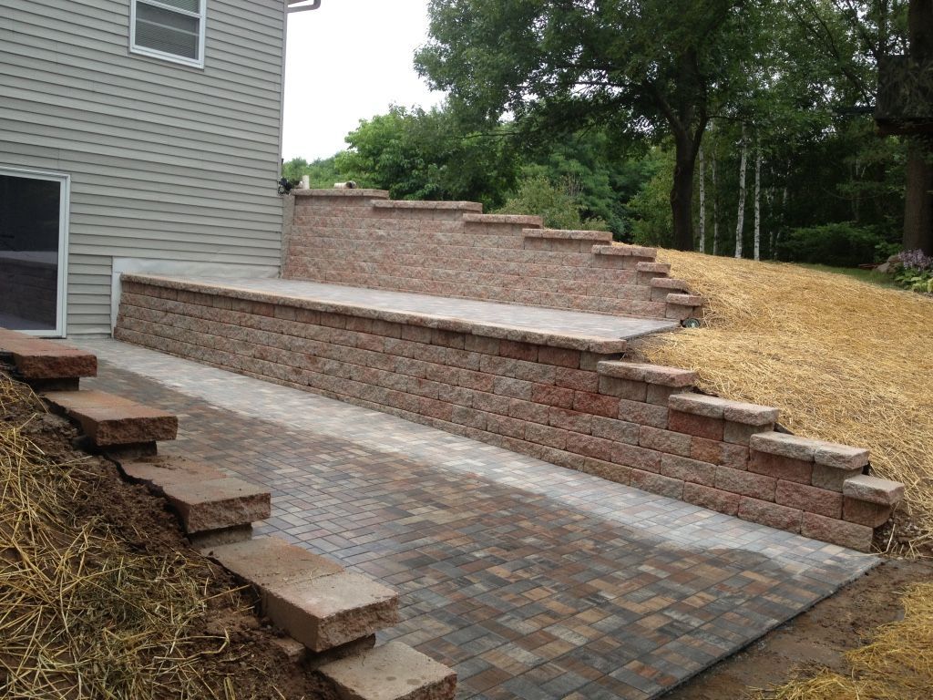 A multi-level stone retaining wall built next to a house, featuring a paver patio and stepped side landscaping.