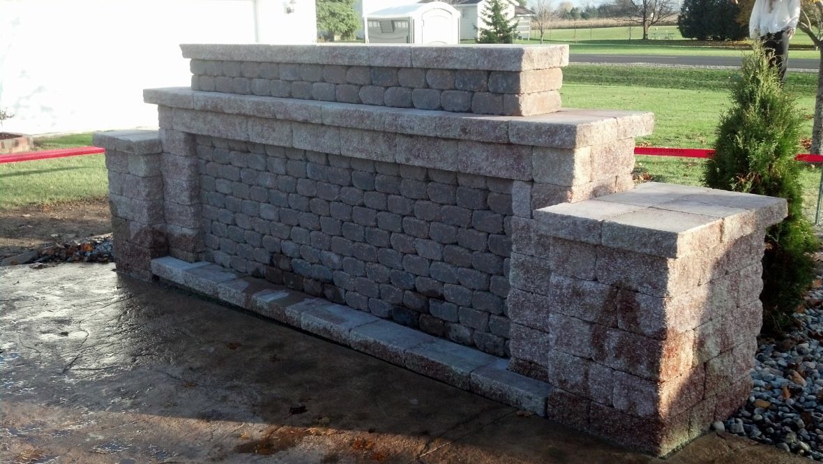 A stone wall feature with tiered, rectangular block construction in a grassy yard, with a small evergreen tree to the right.