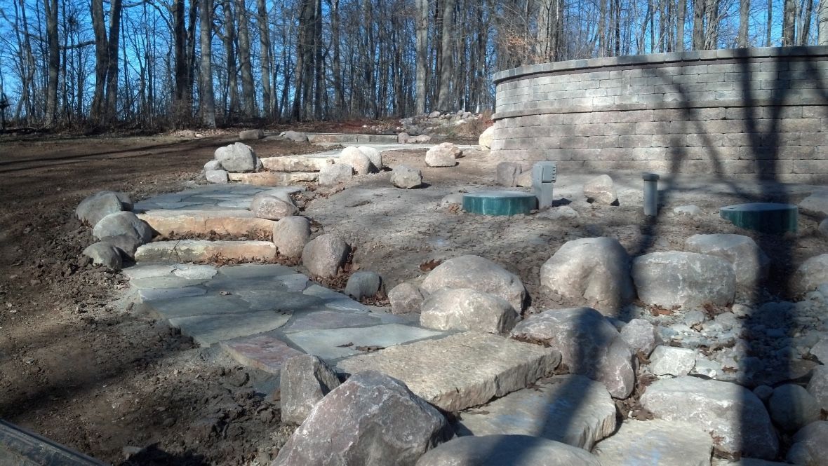 A stone walkway leads to a curved retaining wall in a yard with scattered rocks and a forested background.