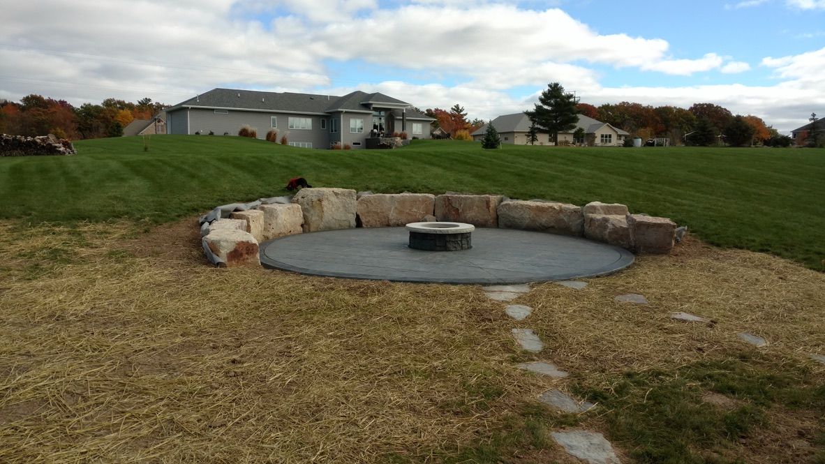 A stone fire pit sits on a circular dark patio, surrounded by a boulder wall in a grassy backyard with homes in the distance.