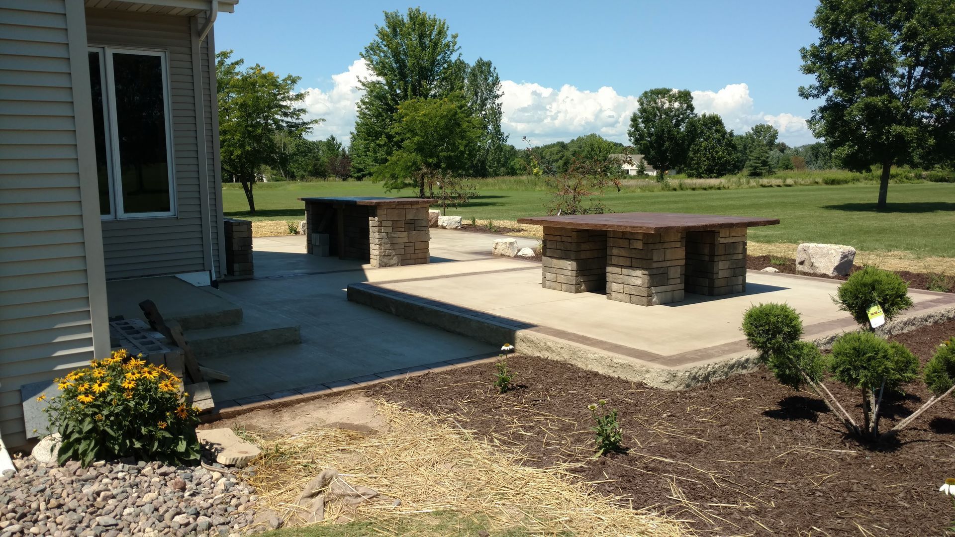 A tan house patio featuring stone-base tables and counters, overlooking a grassy field under a sunny blue sky.