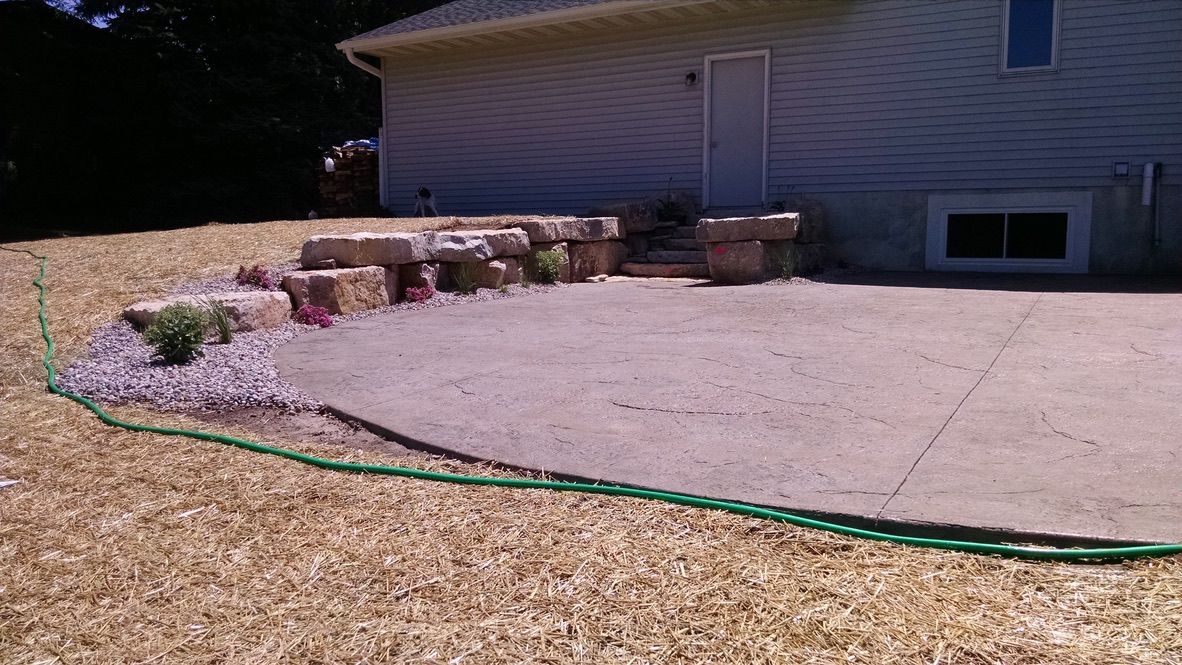 A stone retaining wall curves along a concrete patio next to a house, bordered by a garden bed with rocks and plants.