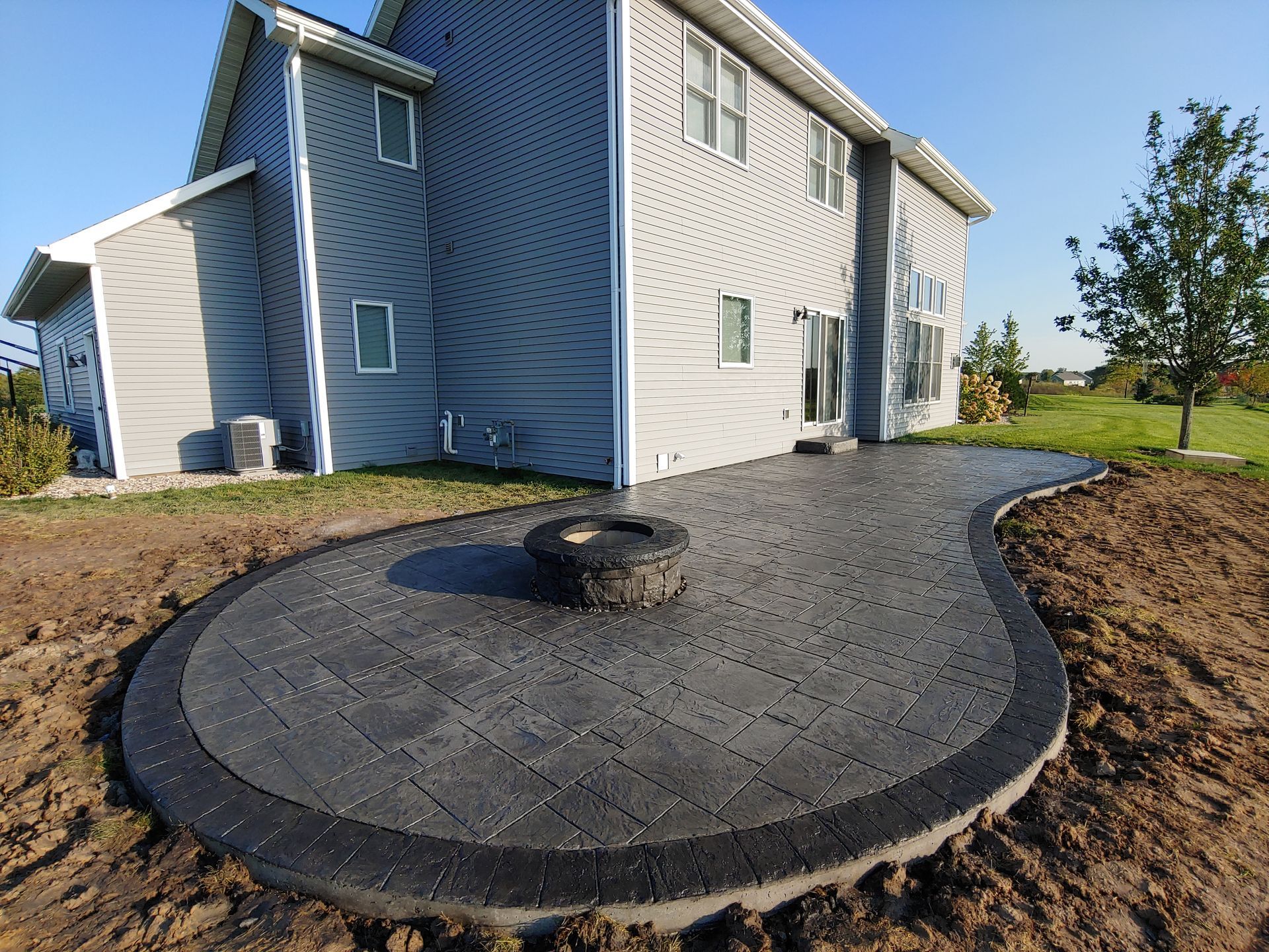 A dark stone patio with a central circular fire pit sits in front of a blue-sided house on a sunny day.