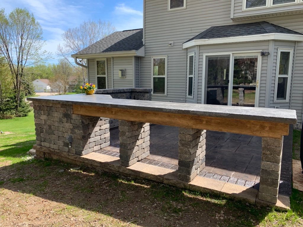 An outdoor stone bar with a wooden countertop on a paved patio in a residential backyard.