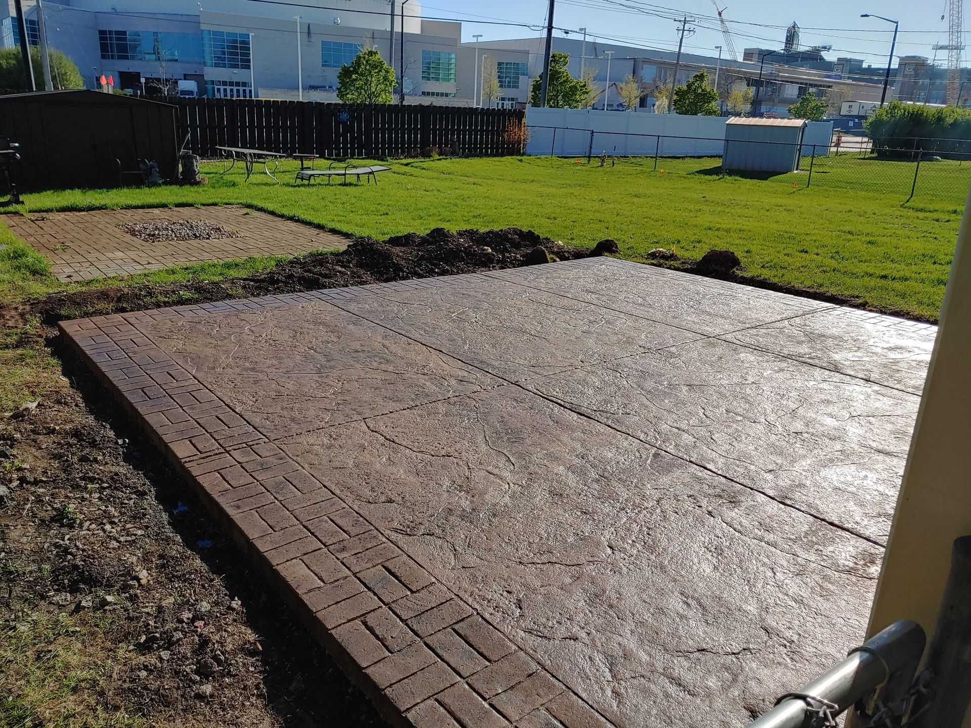 A rectangular paved patio with a decorative border sits in a grassy yard, with a smaller square patch visible nearby.