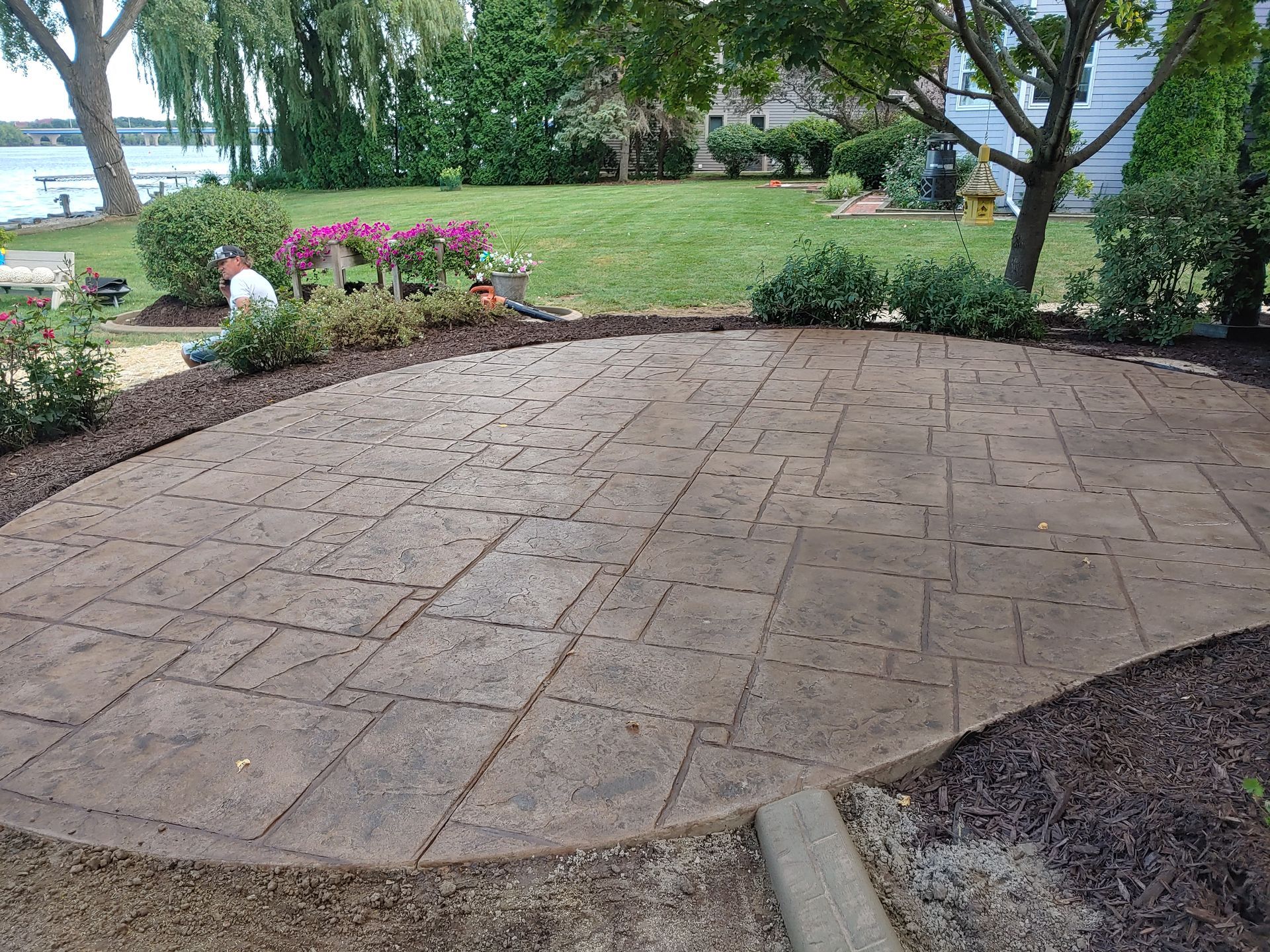A curved, patterned concrete patio sits in a lush green backyard near a lake, surrounded by mulch beds and shrubs.