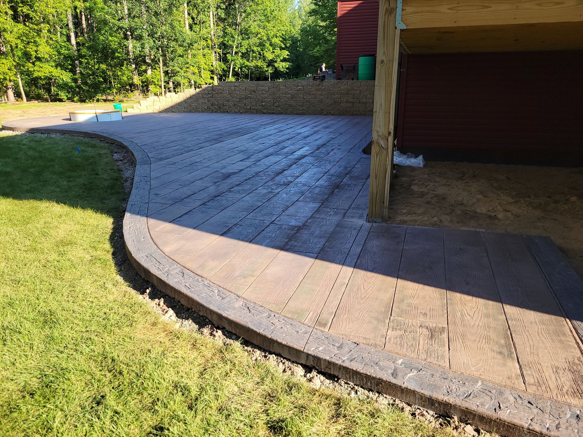 A patio with wood-textured concrete pavers curves along a grassy lawn, extending under a wooden deck support.