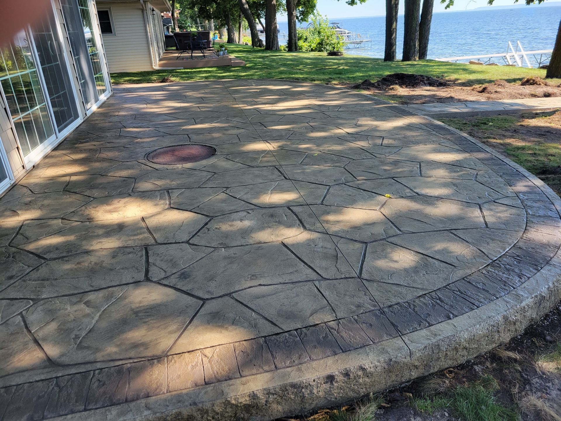 A stamped concrete patio featuring a flagstone pattern with a decorative border, overlooking a lawn and a lake.