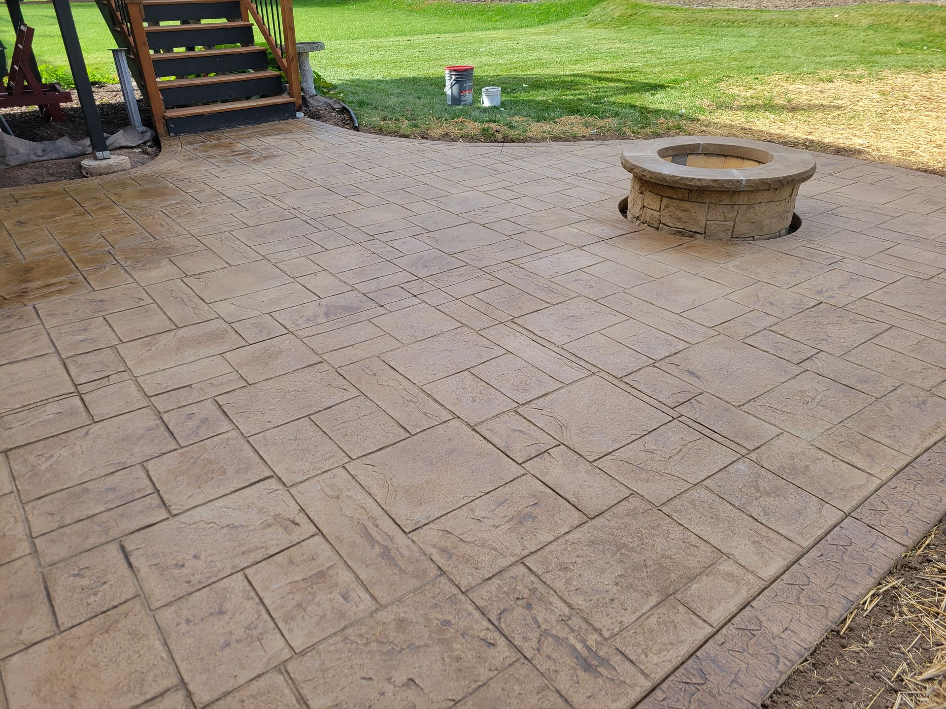 A tan stamped concrete patio featuring a fire pit and adjacent wooden stairs leading to a grassy yard.