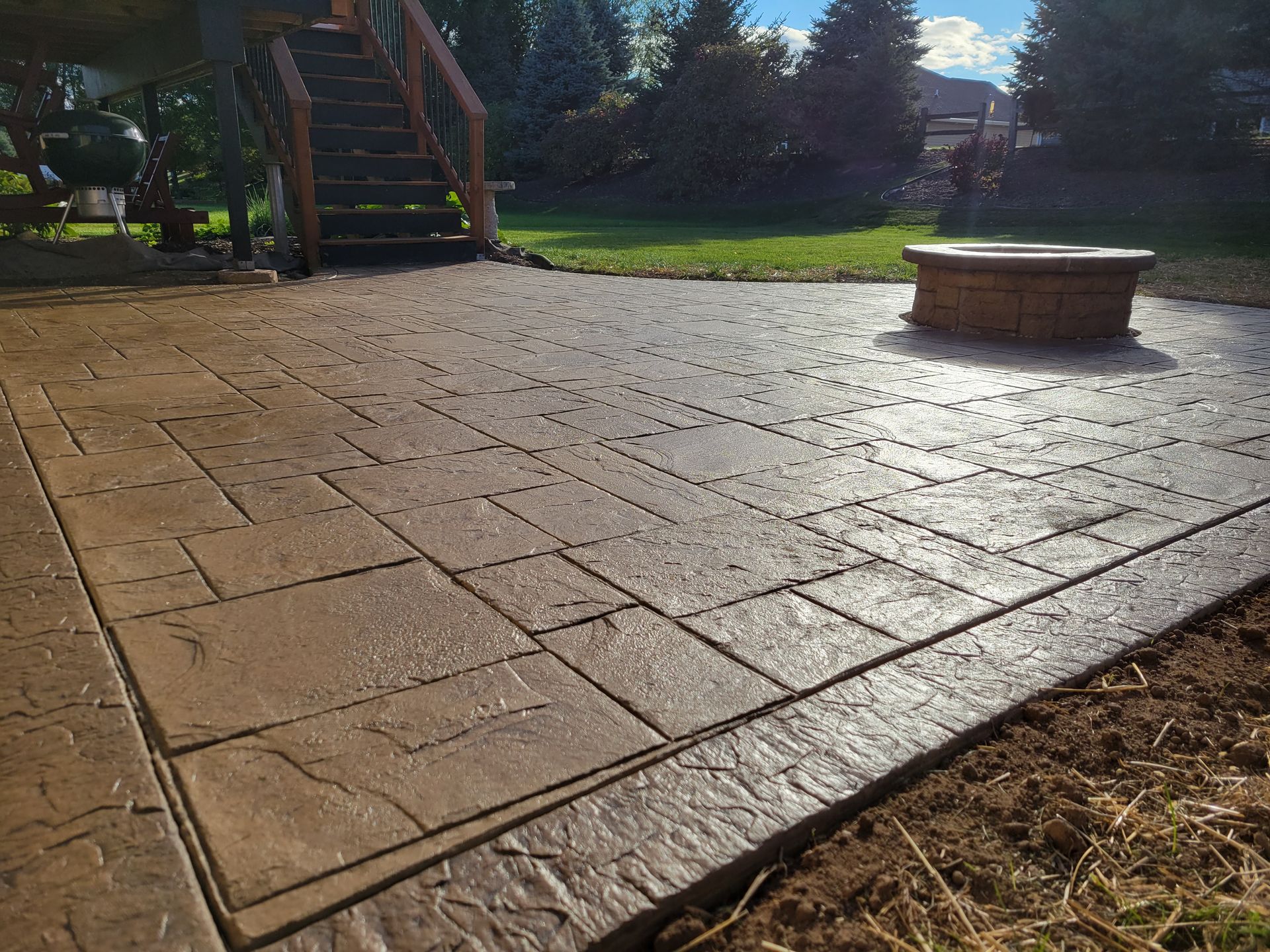 A brown stamped concrete patio features a stone fire pit and wooden stairs leading to a deck in a backyard setting.